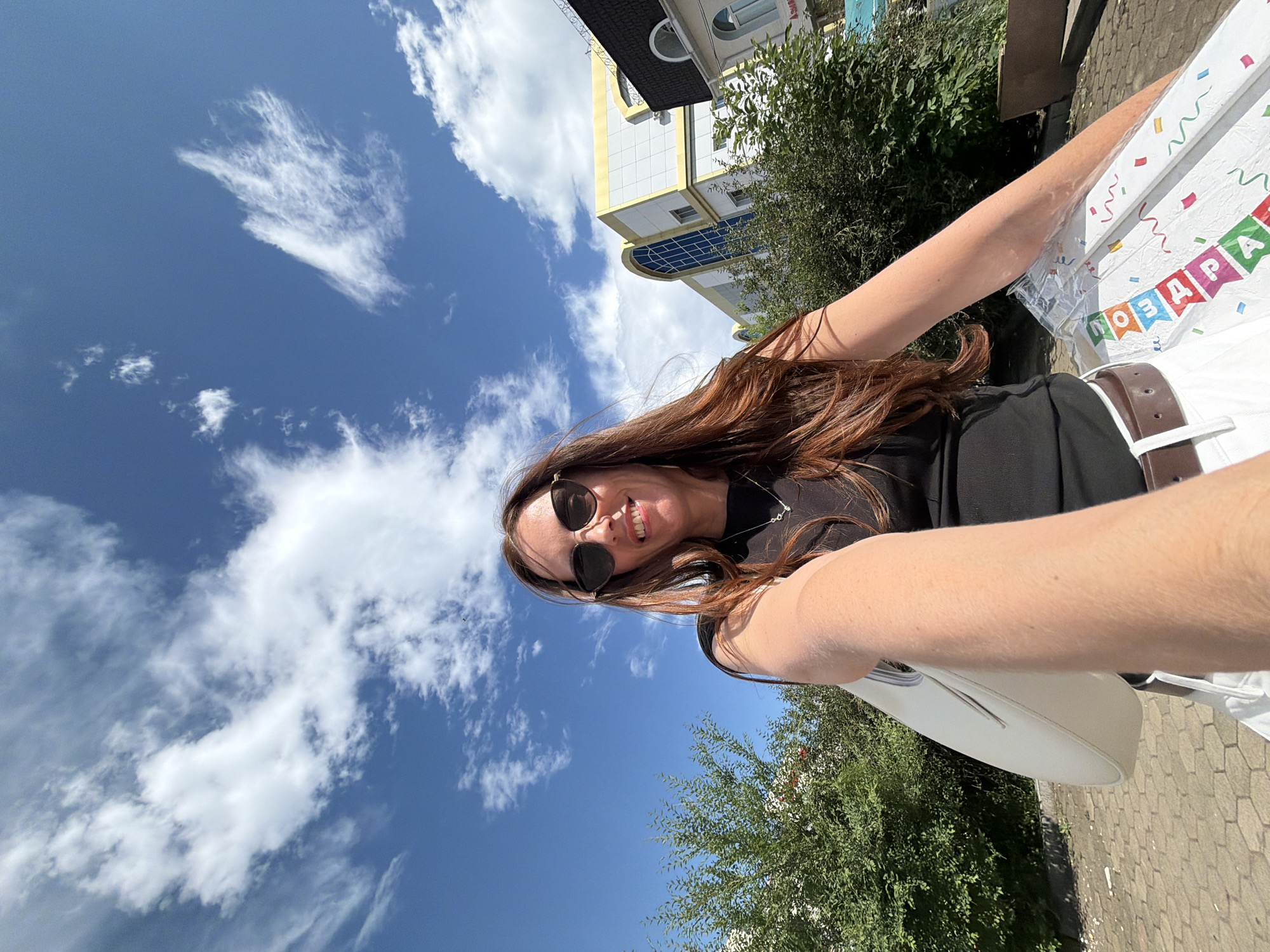 A woman with long, reddish-brown hair and sunglasses smiles at the camera in a selfie. She is wearing a black top, white pants, and a brown belt. She is holding a white purse in her left arm and a package wrapped in plastic with colorful decorations that appear to say "ПОЗДРАВЛЯЮ" (meaning "Congratulations" in Russian) in her right hand. Behind her, there are green bushes, a paved walkway, and buildings with yellow and white facades. The sky is blue with scattered white clouds. The scene appears to be outdoors in Birobidzhan, Russia, under a bright sky.