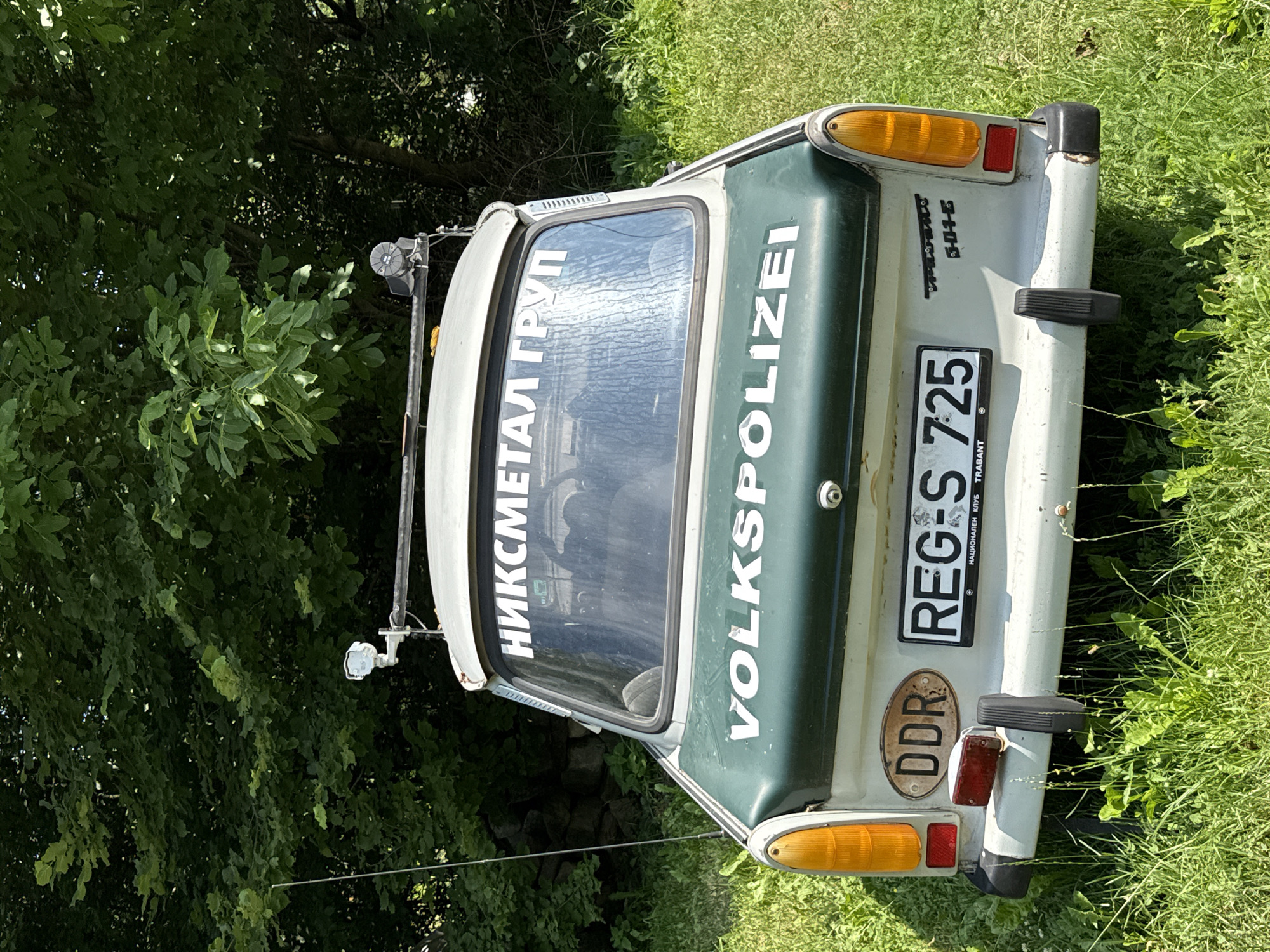A rear view of a light-colored, classic Trabant vehicle, painted green on its rear section, is parked on a grassy area, with lush green foliage forming the background. The car displays several stickers and markings. On the rear window, white lettering reads "НИКСМЕТАЛ ГРУП". Below this, on the green section of the car, are the words "VOLKS POLIZEI" in white capital letters. To the left of the license plate, an oval sticker reads "DDR". The license plate itself is black and white, with the text "REG-S 725". Beneath the license plate, smaller text is visible, including "НАЦИОНАЛЕН КЛУБ" and "TRABANT". On the right side of the rear of the car, a stylized logo with text appears to read "Trabant 601 S". Two orange rear lights are visible, one on each side, along with red reflectors below them. A black bumper extends across the lower rear of the vehicle. The environment is outdoors, with the car situated on uneven ground covered in green grass and some scattered weeds. The image appears to have been taken during daylight. The car is not in motion. No people are visible in the frame. The scene is located in the city of Ribaritsa, Bulgaria.