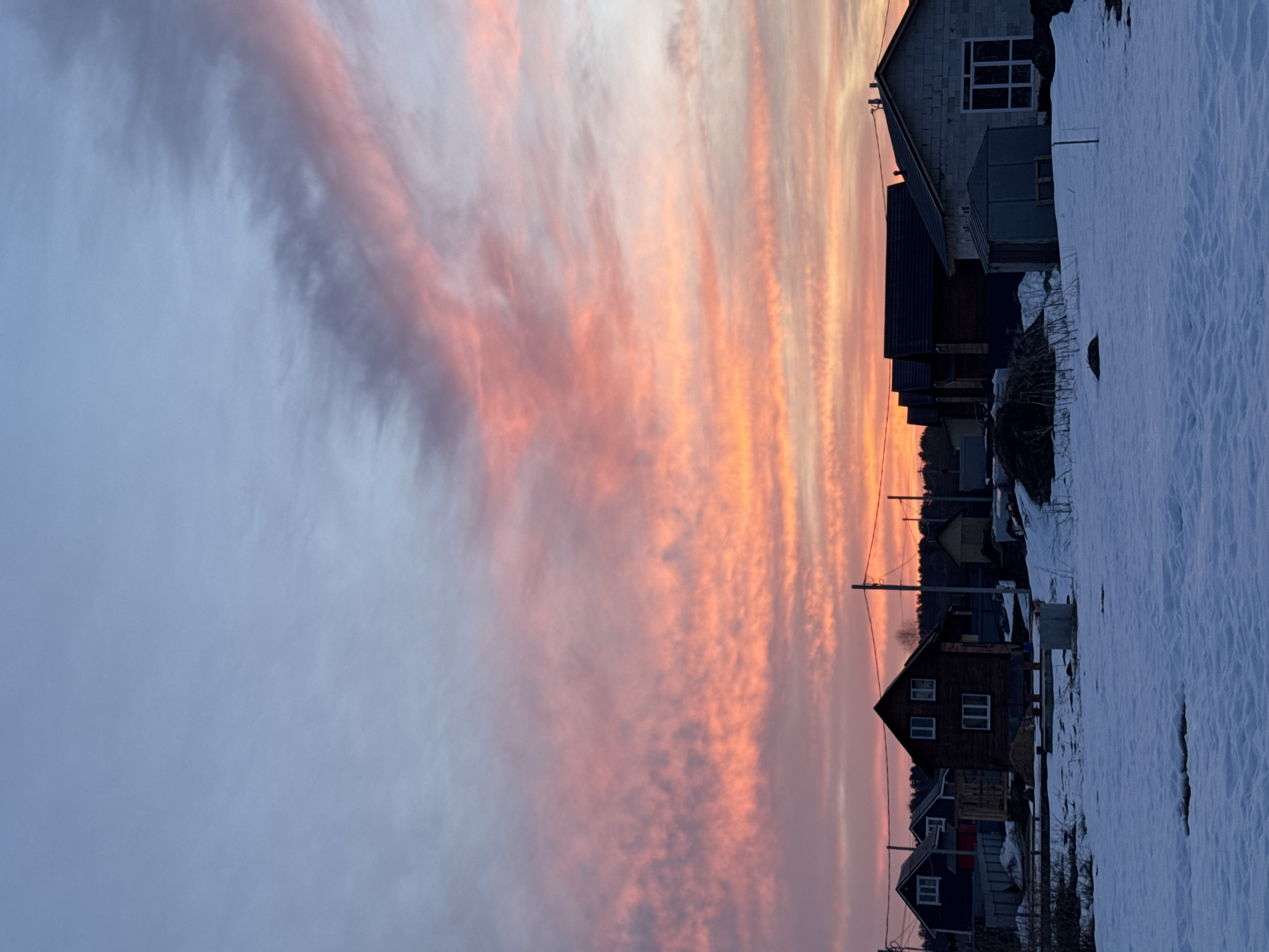 The image captures a row of houses situated along a snow-covered ground in Gornyi, Russia (Dvurechenskoe S/P). The sky above exhibits an array of clouds with varying hues of orange, pink, and gray, likely reflecting a sunrise or sunset. The houses appear to be diverse in style and construction materials. Overhead power lines are visible.