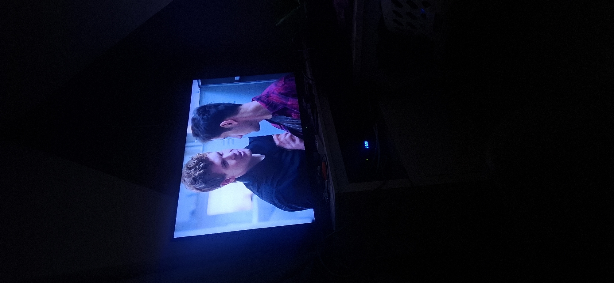 The image shows a television screen displaying a scene with two young men. The man on the left has short, styled blonde hair and is wearing a black shirt. He is looking at the man on the right and appears to be speaking. The man on the right has dark, short hair and is wearing a pink and black plaid shirt. He is looking away from the camera, towards the man on the left. The background of the screen is a light blue, suggesting an indoor setting, possibly a room with white walls or partitions. Below the television, a black electronic device with a blue digital display showing "2320" is visible. The overall scene is dimly lit, with the television screen being the primary light source. The image does not provide any specific information about the city of Yerocham, Israel.