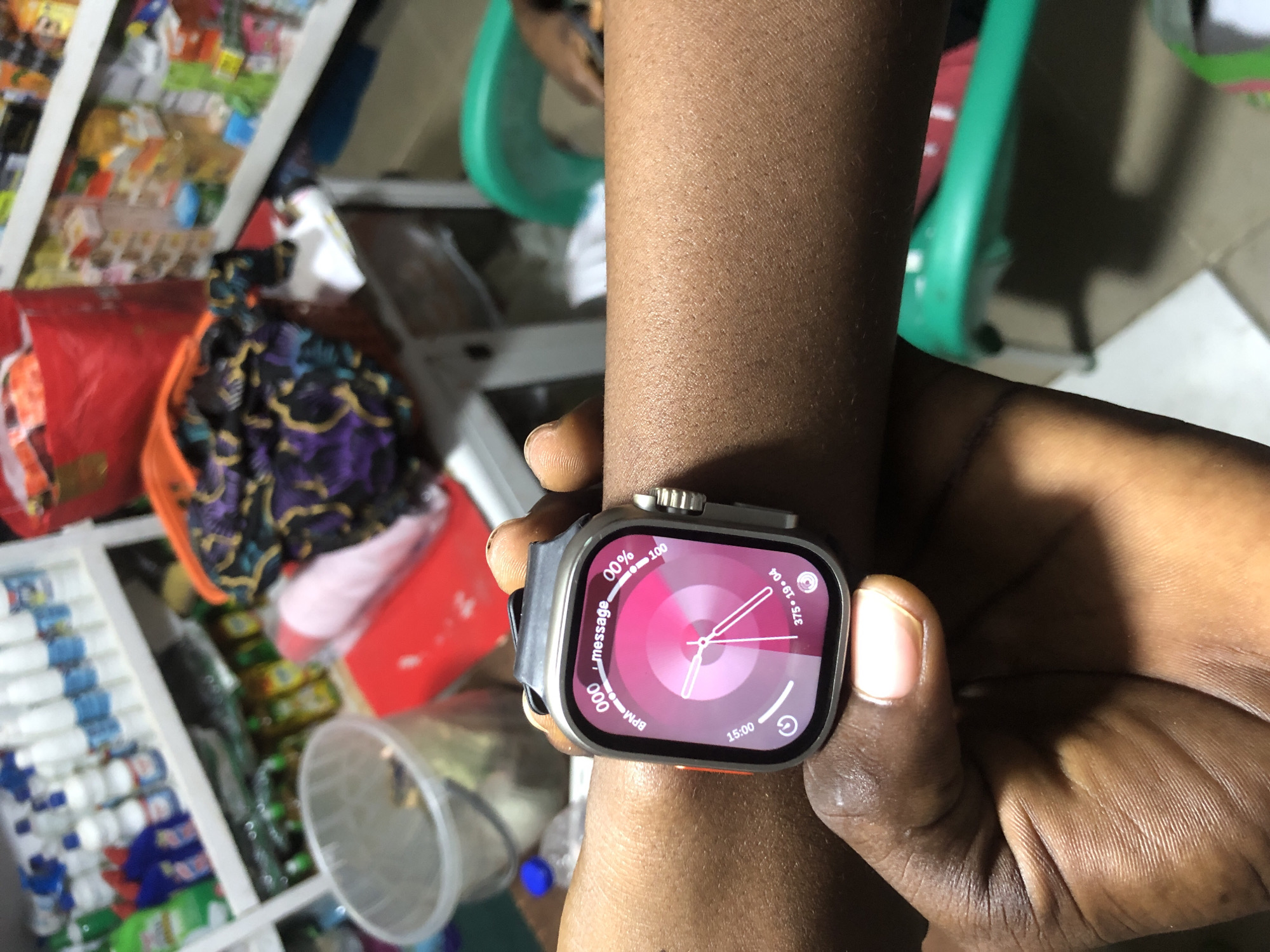 An extreme close-up shot shows a person's left forearm and wrist, which is adorned with a silver smartwatch. The smartwatch's face displays a circular gradient of pink and purple hues, with white text indicating "000 message," "BPM," "15:00," "00% 100," and "375 • 19 • 04." The person's dark-skinned forearm is visible with small pores. The background is blurred but suggests an indoor setting, possibly a retail environment, with shelves stocked with various items, including cleaning supplies and packaged goods. A red plastic bag and some folded fabric are also discernible in the background. The overall scene is well-lit, focusing attention on the smartwatch and the immediate area around the person's arm. The image was captured in Ado, Nigeria.