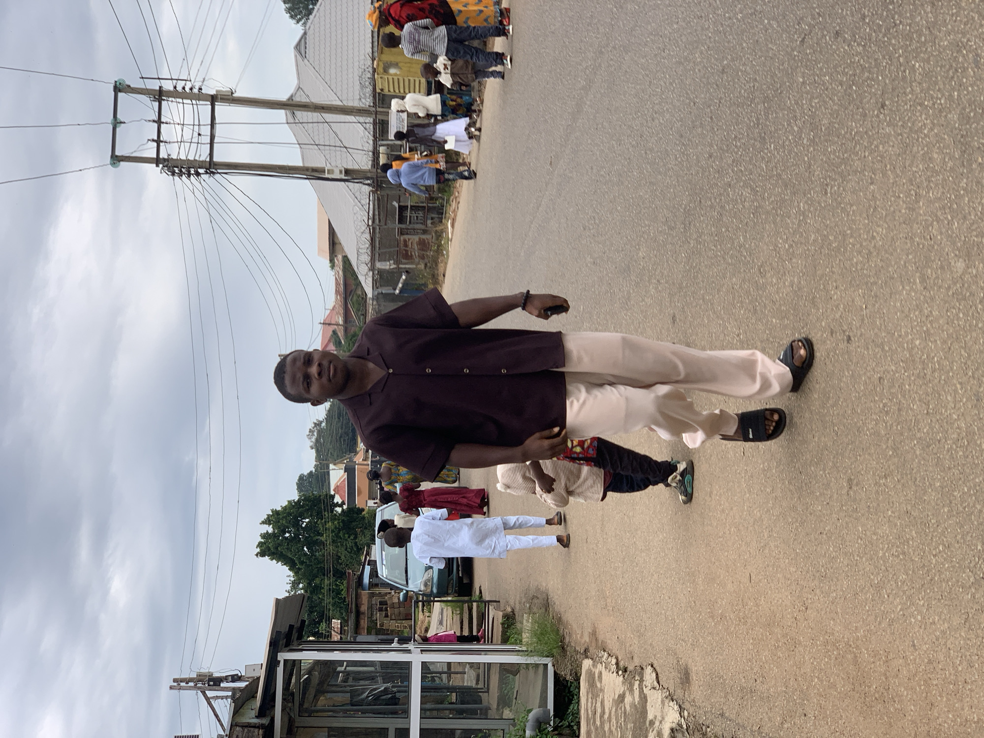A medium shot captures a street scene in Jos, Nigeria, under an overcast sky. In the foreground, a young Black man walks toward the camera, wearing a dark brown short-sleeved collared shirt and light beige pants. He carries a dark object, possibly a phone, in his right hand. To his left and slightly behind him, a young Black child in a hooded outfit walks away from the camera, while another child in dark pants and a striped shirt is partially visible.
Further down the street, several other individuals are present. A man in a white traditional outfit walks away from the camera, followed by a woman in a red dress. In the background, a light-colored car is parked on the side of the road. Other people, including women and children, are seen walking and standing near buildings and a large utility pole with numerous wires. The street is paved with asphalt, and the sides of the road are lined with buildings, some with shops, and vegetation. The overall atmosphere appears to be a typical day in a residential or commercial area of Jos.