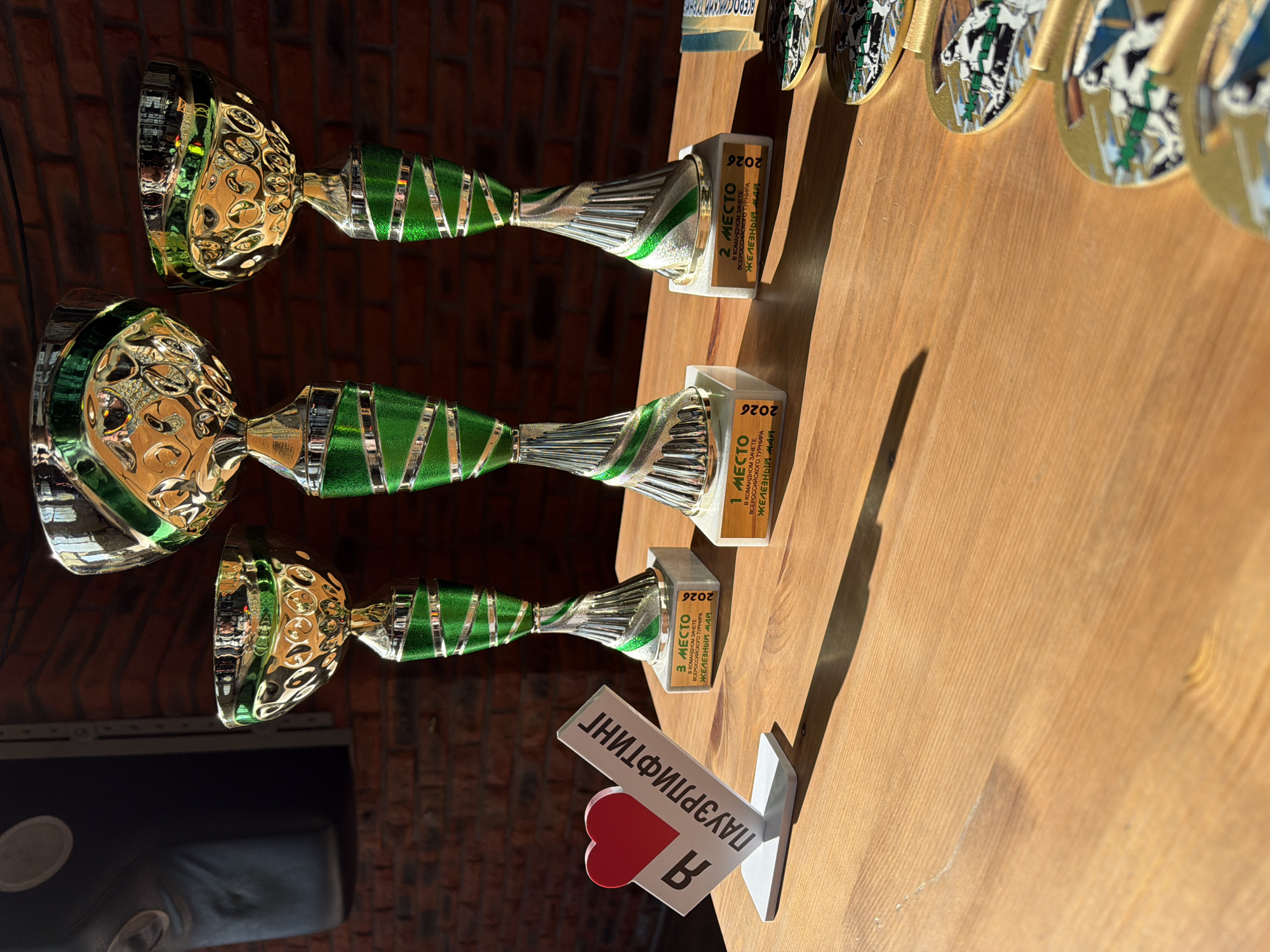 The image shows three trophies and a small sign on a light brown wooden surface. The trophies are arranged in a row, with the bases displaying "3 MECTO" (3rd place), "1 MECTO" (1st place), and "2 MECTO" (2nd place), respectively. Each base also contains Russian text about a team event in the "Железный май" (Iron May) All-Russian tournament, held in 2026. The trophies are silver and green with a golden cup. Next to the 3rd place trophy is a white sign with red heart and Russian text reading "В ПАУЭРЛИФТИНГ". In the upper right of the frame are a series of gold-colored medallions.
The background behind the trophies appears to be a brick wall and a dark object, possibly a leather seat.
The scene is likely related to a powerlifting competition or event in Chaikovsky, Russia given the Russian text and trophies.