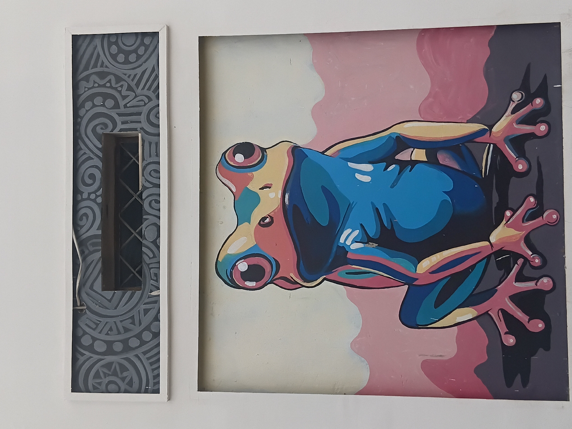 A brightly colored painting of a frog hangs on a wall next to a decorative panel with a dark gray and white pattern. The frog is depicted in shades of blue, pink, yellow, and black against a painted background of light pink and gray. The decorative panel features a vertical rectangle opening with a gray grill. The overall setting is stark white. No information on the location of Gura Top, Nigeria is evident from the image.
