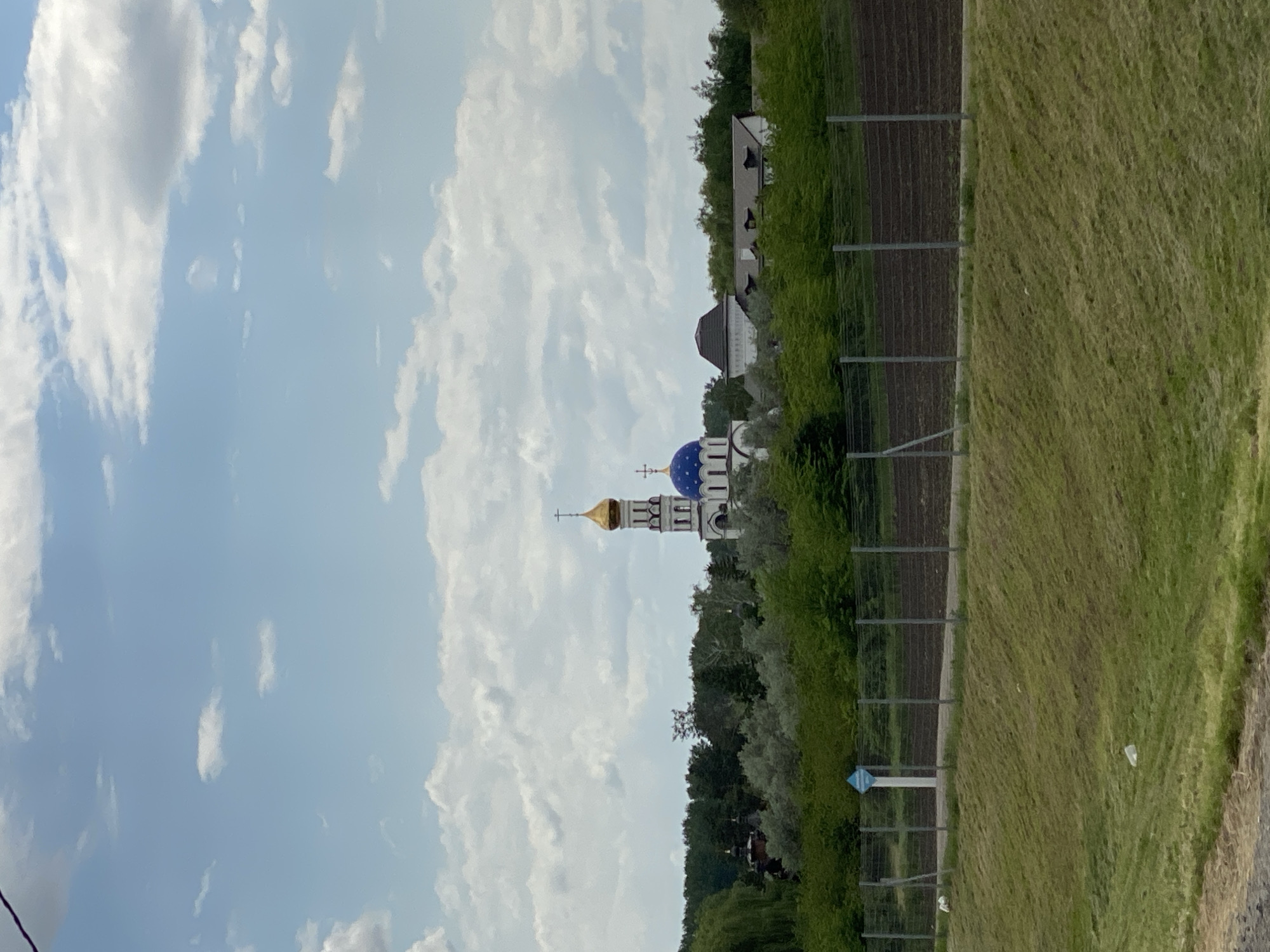 The image shows a landscape with a church in the background. The church is Orthodox with a blue and gold dome. There is a field and a fence in the foreground. The sky is cloudy. The location is Plastunovskaya, Russia.
