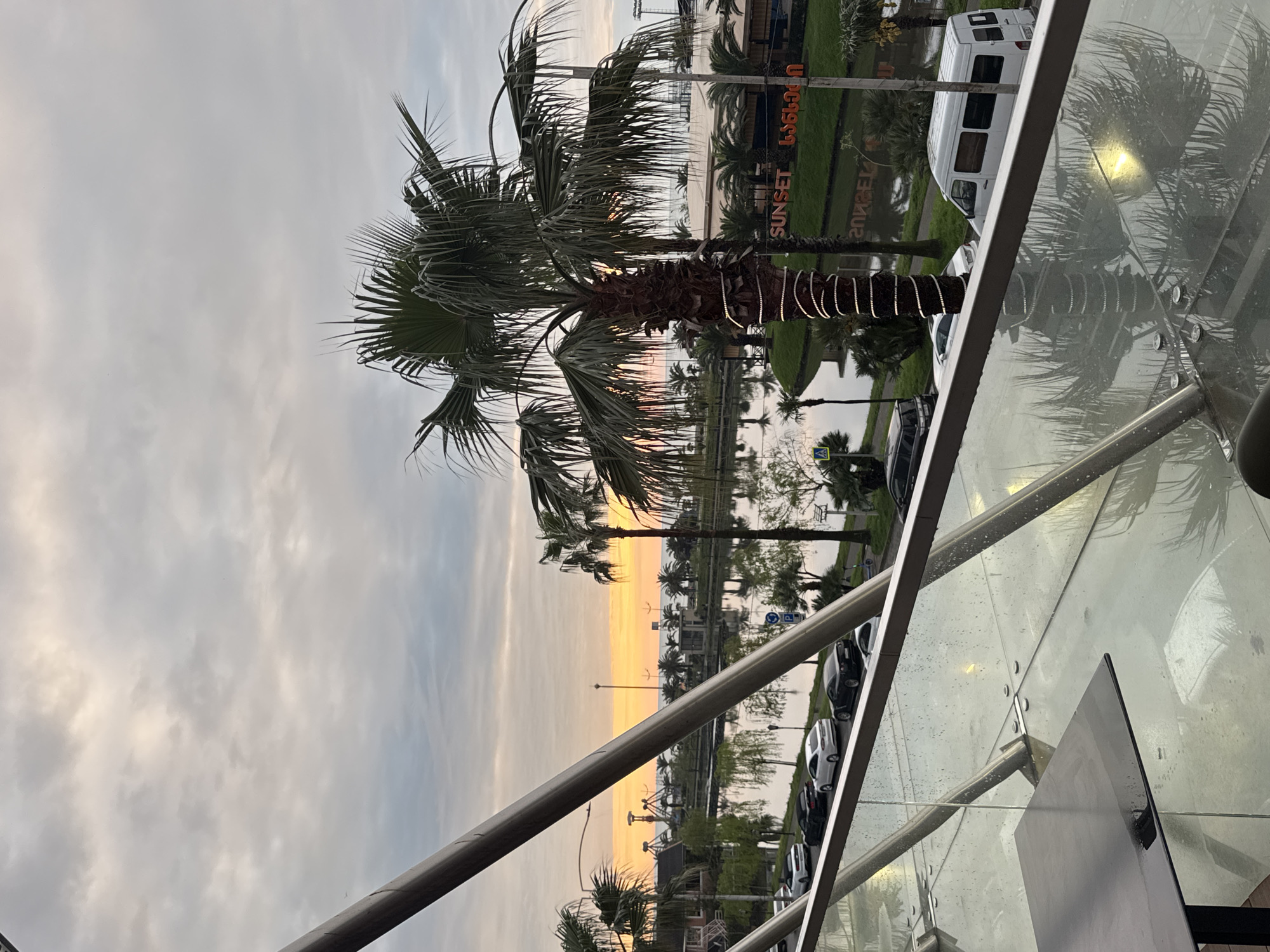 Captured from an elevated viewpoint, the image shows a sunset scene in Batumi, Georgia. Prominent in the frame is a palm tree, adorned with decorative lights wrapped around its trunk. The background reveals a waterfront area with the word "SUNSET" displayed in large letters, possibly indicating a restaurant or entertainment venue. A white van and several cars are parked nearby. The sky displays a blend of colors from the setting sun, visible through the palm tree fronds. A clear glass railing with visible rivets runs along the bottom right of the image, with water droplets suggesting recent rain. A partial view of a table is present in the lower right corner.