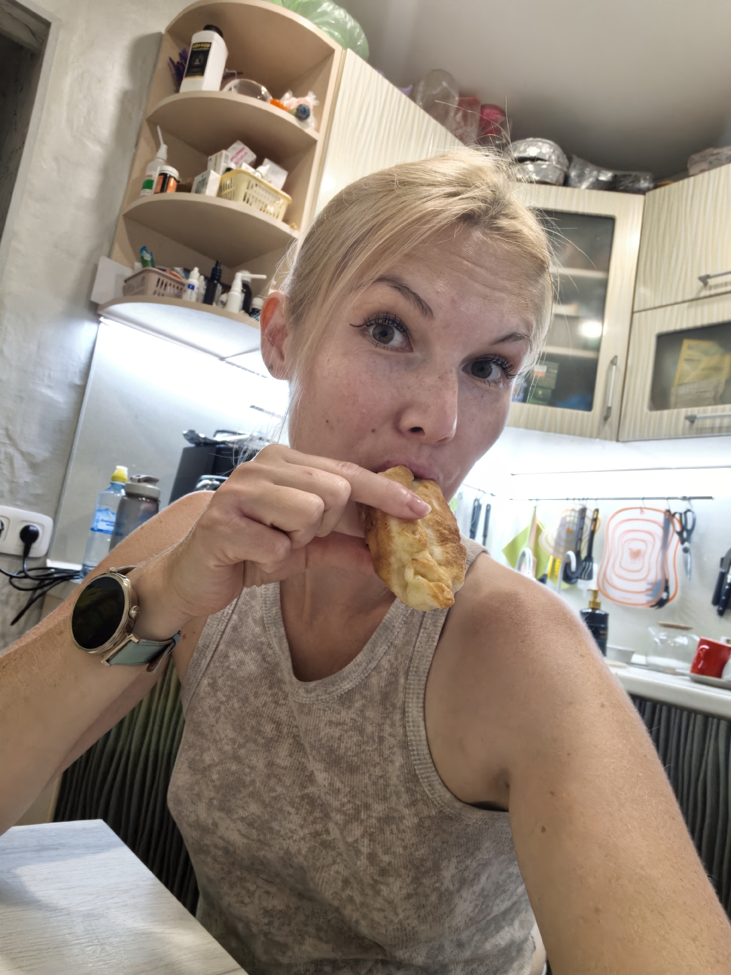 A medium-shot, selfie-style photograph shows a blonde woman with fair skin and freckles in a kitchen. She is wearing a light grey, sleeveless tank top and a round smartwatch with a light blue band on her left wrist. Her right hand is bringing a piece of golden-brown fried dough, possibly a pastry or pierogi, to her mouth. Her eyes are open and looking at the camera, and her mouth is slightly open as she is about to take a bite.
The background depicts a kitchen environment. On the left, a white electrical outlet with a black plug is visible on a light grey wall. To the right of the outlet, a clear plastic water bottle with a blue label and a grey and black water bottle are on a counter. Above the counter, a light-colored shelf unit with rounded corners holds various items, including bottles, containers, and what appear to be toiletries or medicines.
Further to the right, kitchen cabinets with glass-fronted sections are visible. Below the cabinets, a countertop features several items, including a red mug, a black bottle with a pump dispenser, and kitchen utensils hanging on a rack. The overall lighting in the kitchen appears to be artificial, with illuminated strip lights under the cabinets. The scene suggests a casual moment in a domestic setting. There is no information about the specific location within the city of Sterlitamak, Russia.