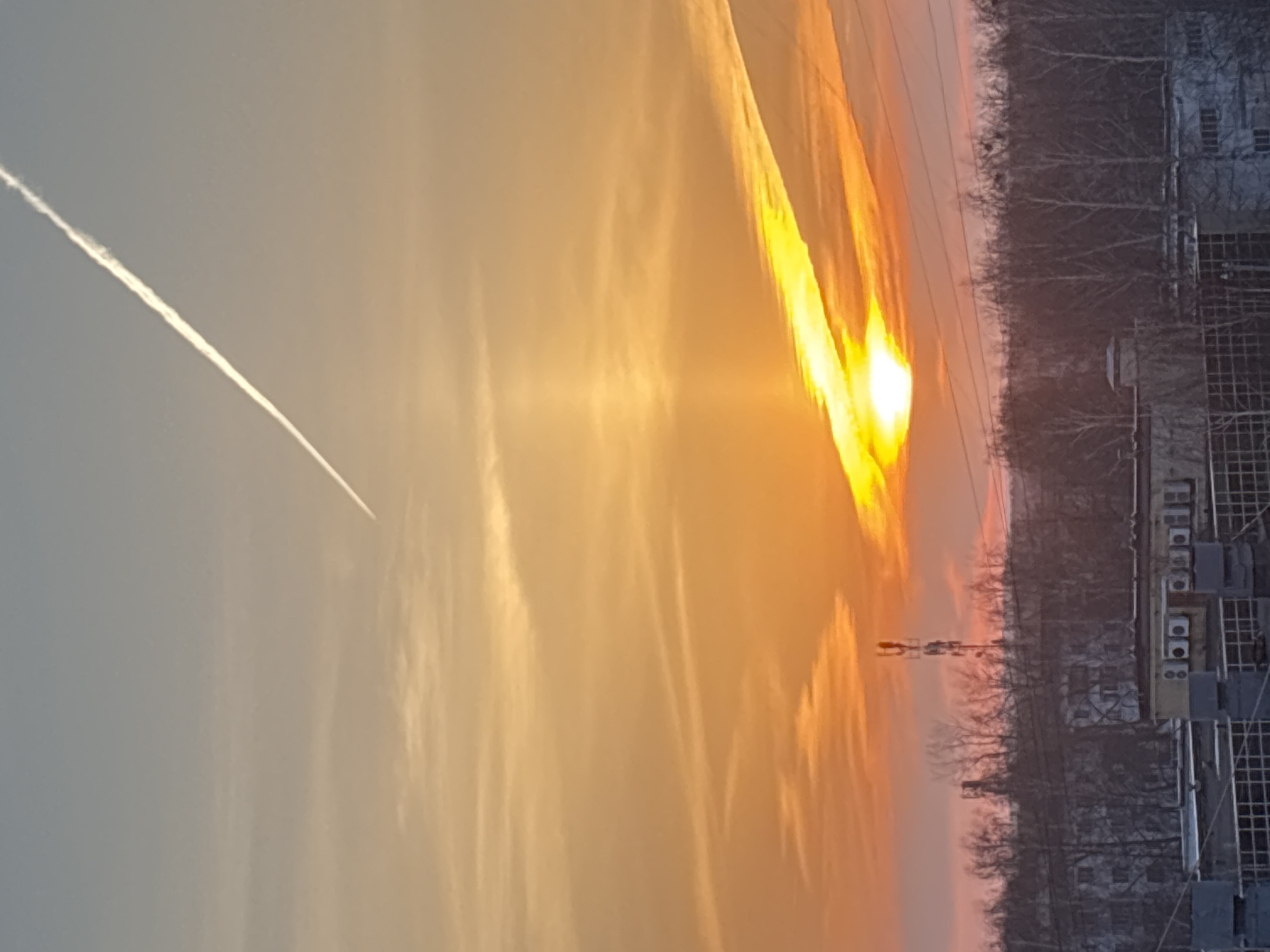 The image shows a sunset over Chaikovsky, Russia. A vapor trail from a plane is visible in the sky. The sky is filled with clouds, varying in hues from light grey to orange. The sun is near the horizon, with bright yellow light streaming through a gap in the clouds. In the foreground, the outlines of a building and a treeline are visible. An electrical pylon is also visible against the backdrop of the sunset. The photo is taken from a low angle.