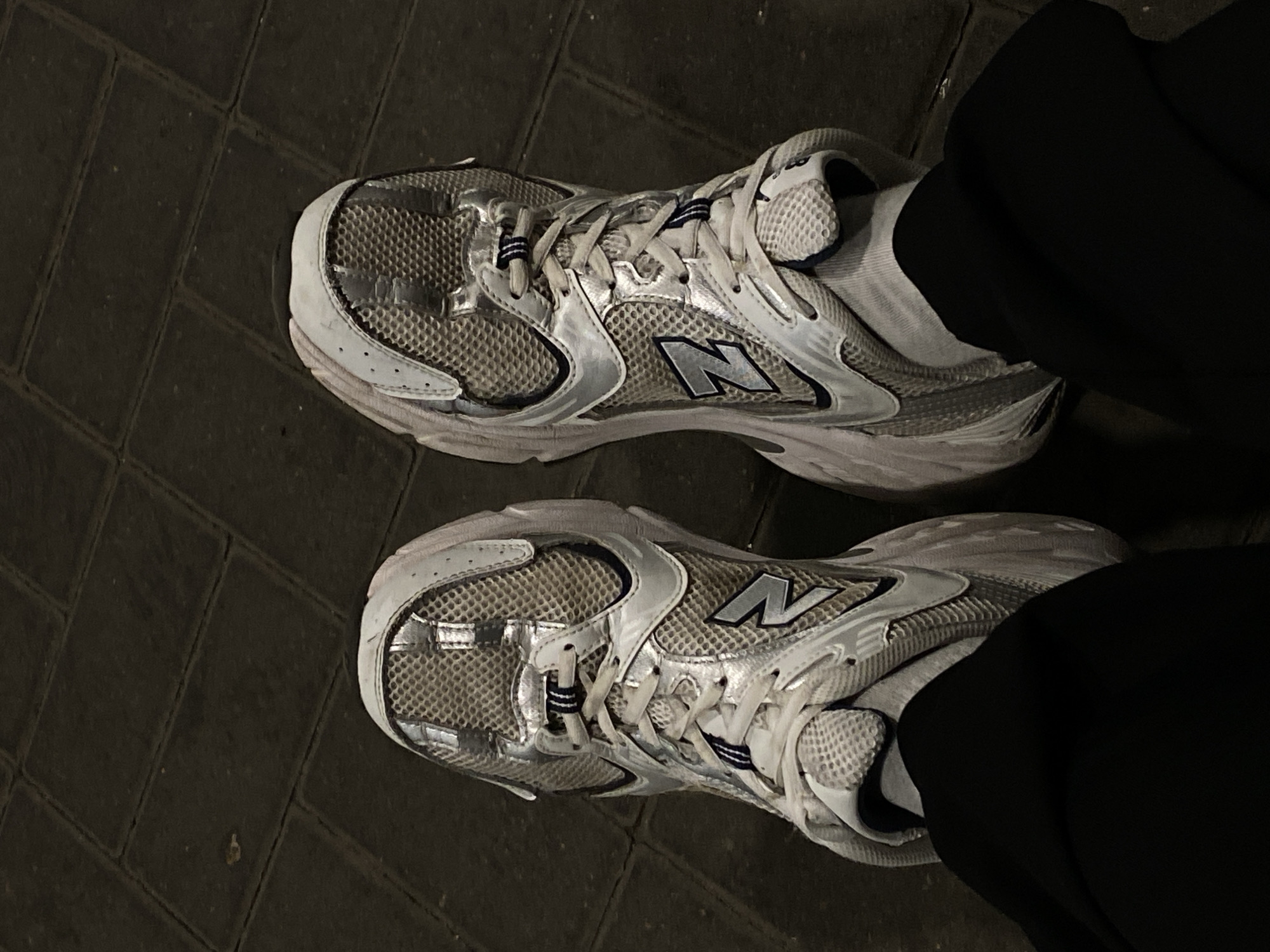 A top-down shot displays a pair of gray and silver New Balance running shoes. The shoes are positioned on a dark gray brick surface, likely a sidewalk or pedestrian area. The shoes are worn with white socks and the legs are clad in dark, likely black, fabric. The scene appears to be dimly lit. There is no information to suggest it is located in Saint Petersburg, Russia.