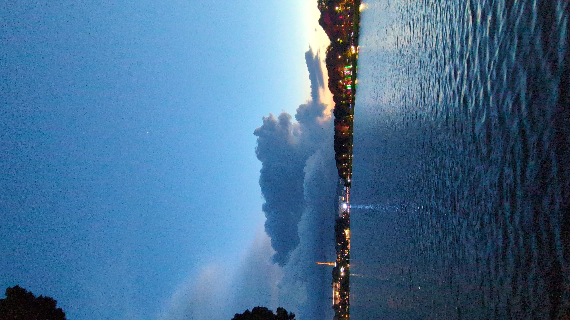 The image captures a serene evening scene at Xuan Huong Lake in Da Lat, Vietnam. The sky is a deep blue, transitioning to hues of orange and yellow near the horizon, suggesting either dawn or dusk. Dark, dramatic clouds are visible in the upper left portion of the sky. Across the lake, the city lights twinkle along the treeline, illuminating the area with vibrant colors, especially noticeable in the cluster of lights towards the right. A prominent, illuminated tower stands out on the left side of the cityscape. The surface of the lake is calm, with gentle ripples reflecting the ambient light and creating a shimmering effect. A dark silhouette of a tree branch is visible in the upper left corner, framing the scene. No people are discernible in the image.