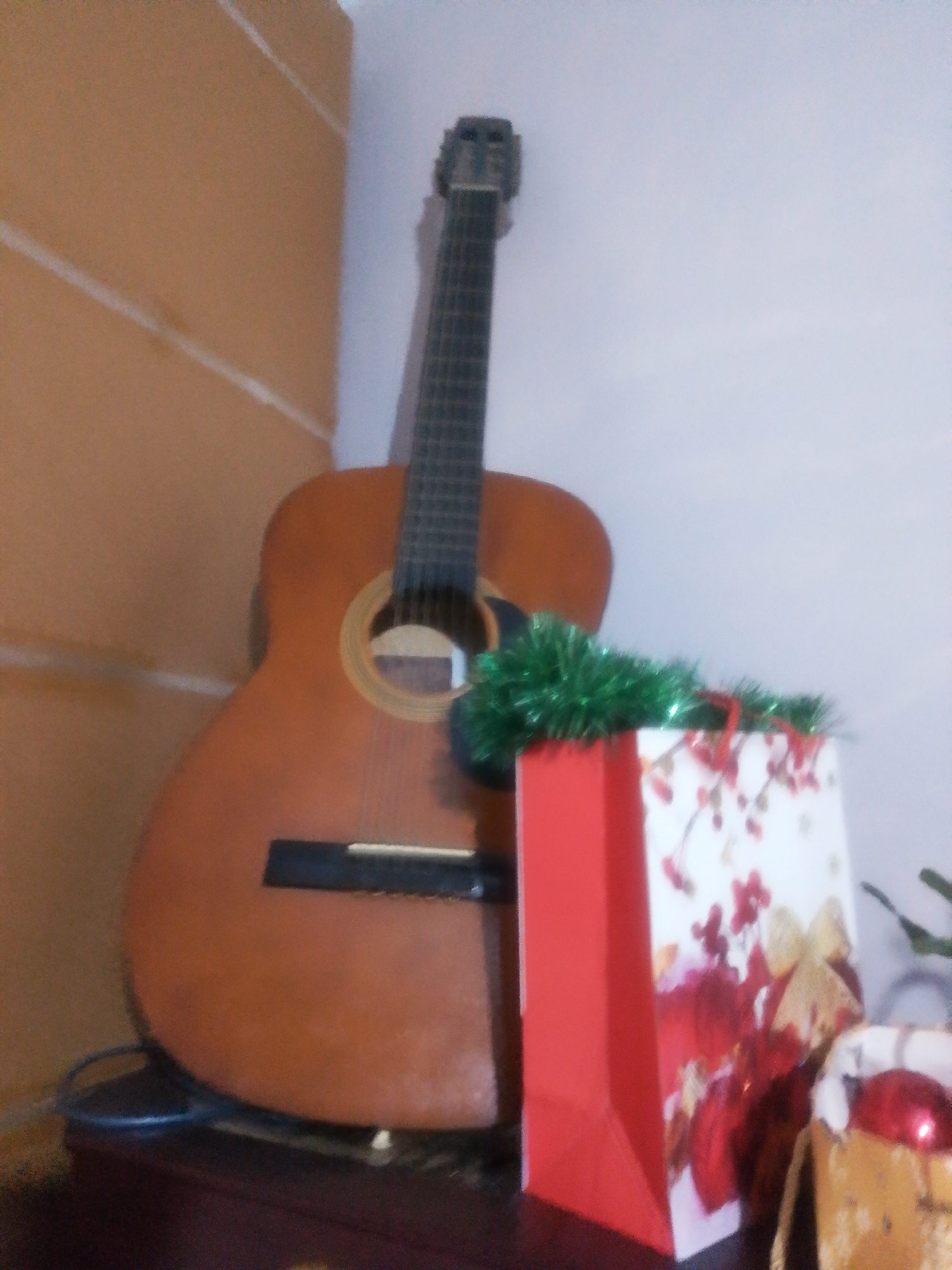The image shows an acoustic guitar leaning against a wall with horizontal paneling. In front of the guitar, a red gift bag and a white gift bag with a floral pattern are placed on a dark surface. Green tinsel is draped over the gift bags. A portion of what appears to be another gift bag with a red ornament is visible on the right side. The scene is indoors, possibly in a domestic setting. There are no people visible in the image. The image is slightly out of focus and grainy. The location in the city of Tekeli, Kazakhstan, is not discernable from the image content.