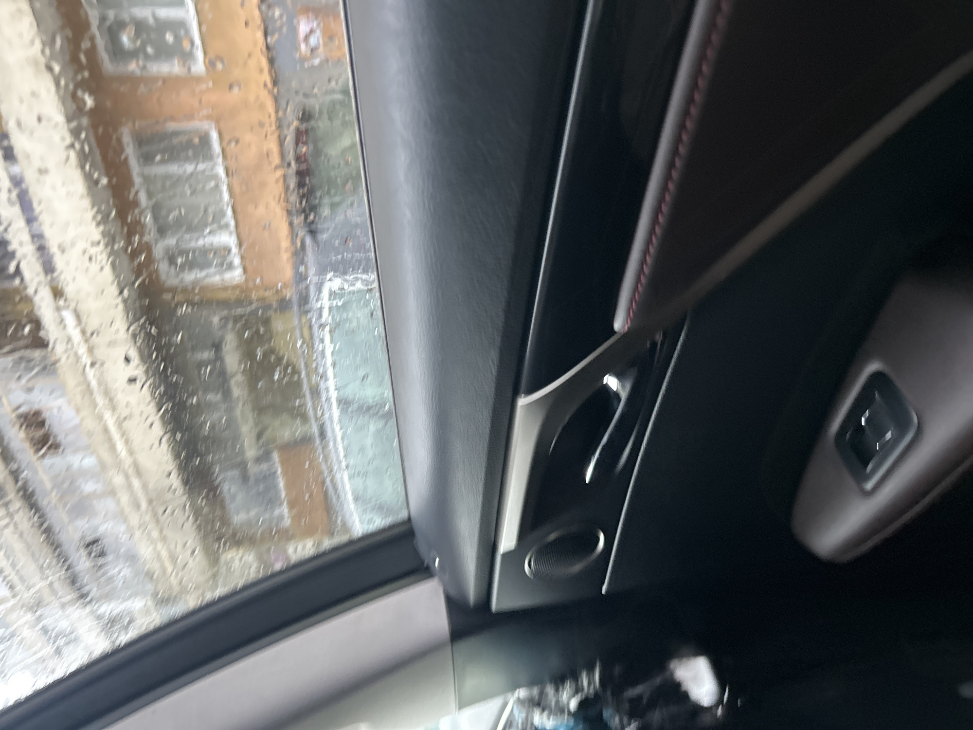 The image shows the interior of a car, possibly parked or stationary, as evidenced by the rain-streaked window revealing a blurred view of buildings. The building facade visible through the window suggests an urban setting with structures of light-colored brick or stucco. The car's interior features dark-colored trim, an air vent, and a seat adjustment control. The location cannot be determined from this image alone.