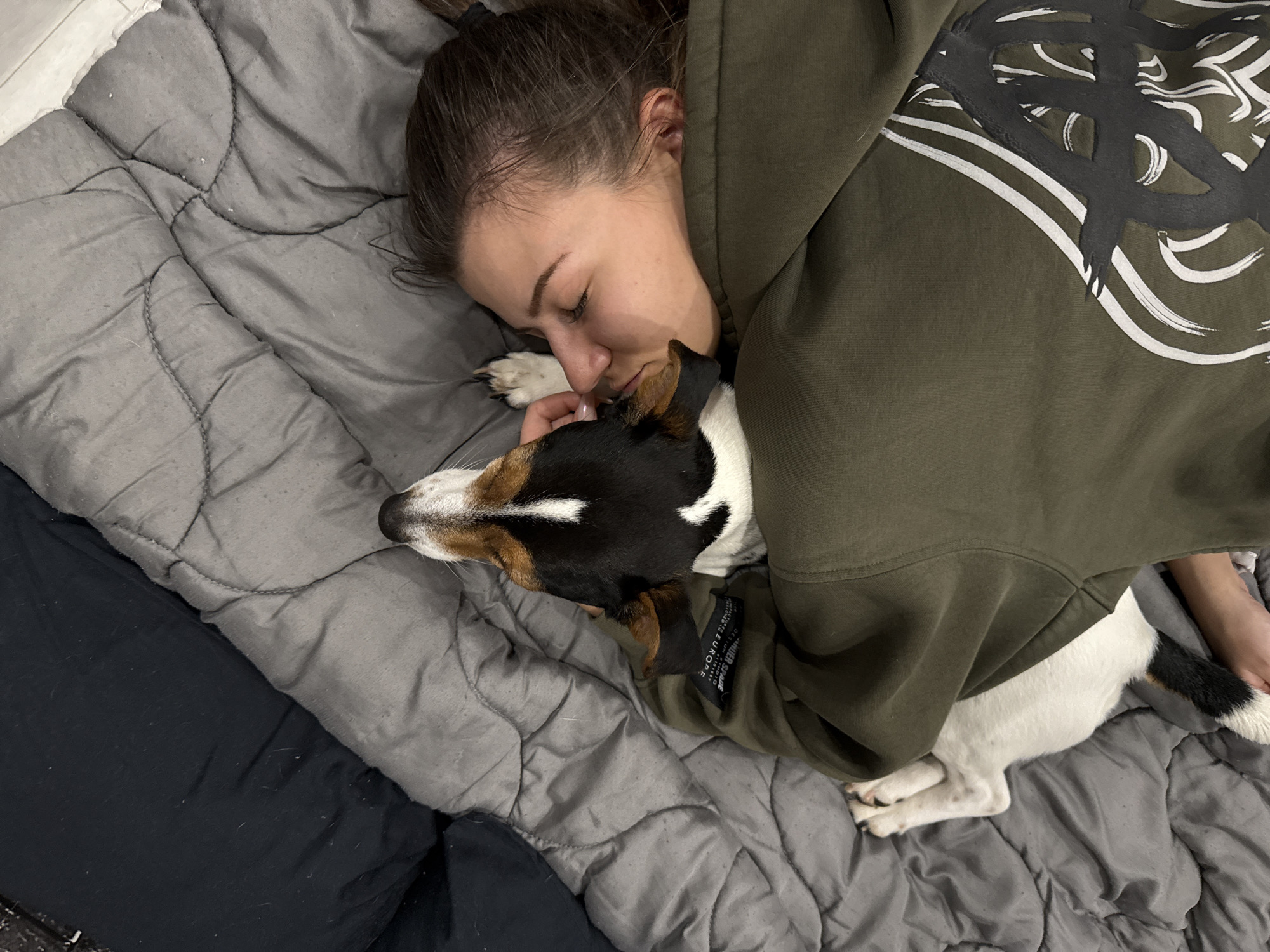 A young woman lies in bed with a Jack Russell Terrier. She is lying on her side facing the dog with her eyes closed. She is wearing an olive green hoodie with a white and black design. The dog is lying next to her with its head resting on her chest. The woman's right arm cradles the dog's head. The bed has a gray duvet. The location is inside an apartment or house; there are no indicators the image was taken in Sofia, Bulgaria.