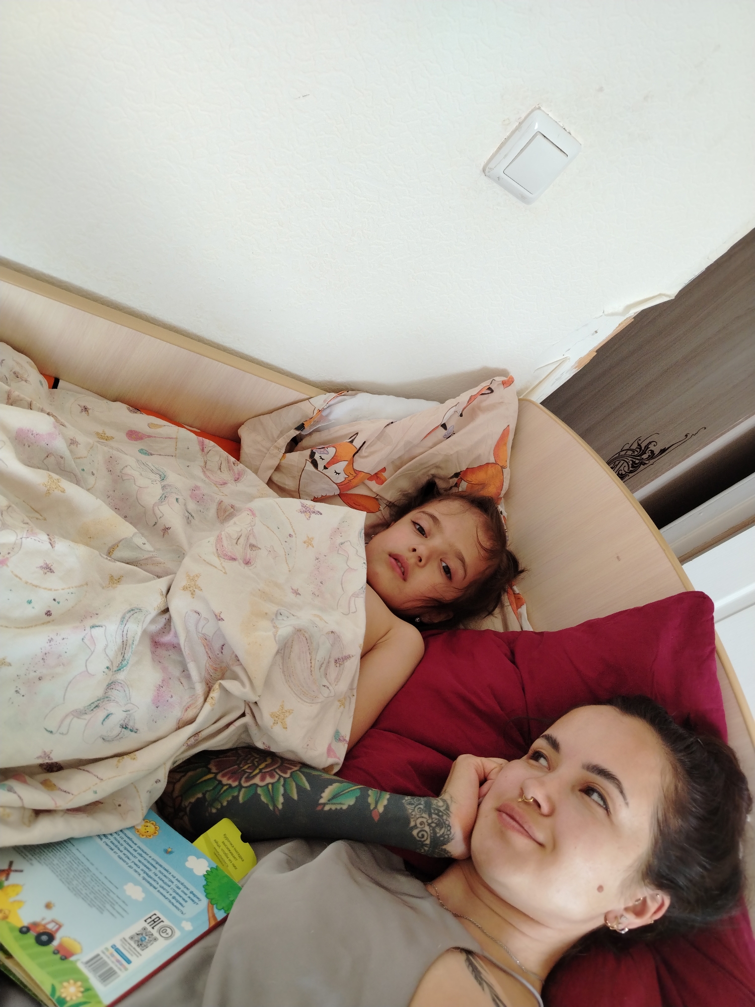 A woman and a young child are lying in a bed. The child is on the left side, covered with a blanket. The woman is on the right side, resting her head on her hand. A book lies beside them. A light switch is on the ceiling. Location is Noviy, Russia.