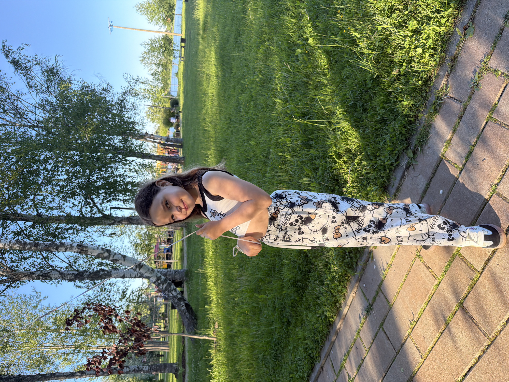 This image depicts a young girl standing outdoors on a sunny day in a park-like setting in Zaprudnya, Russia. The girl, appearing to be 6-10 years old, has dark brown hair and is looking towards the camera with a slight smile. She is wearing a white sleeveless crop top with black trim, featuring "Hello Kitty" graphics, exposing her midriff. Her pants are loose-fitting, white with a black and brown "Hello Kitty" graffiti-style print. She wears white and black sneakers with a light brown sole. In her left hand, she holds a thin, silver-colored string, likely from a balloon extending out of frame upwards. She stands on a light-colored paved path composed of rectangular blocks, with vibrant green grass to her right. The scene is brightly illuminated by direct sunlight, casting distinct shadows from the girl and unseen objects onto the path and grass. In the background, numerous deciduous trees, including several prominent birch trees with white bark, are visible. Beyond the trees, a clear blue body of water, likely a lake or river, stretches under a bright blue sky. Distant figures of people and some indistinct structures, possibly part of a playground or recreational area, are visible across the water or further within the park. A wooden utility pole stands erect near the distant water.