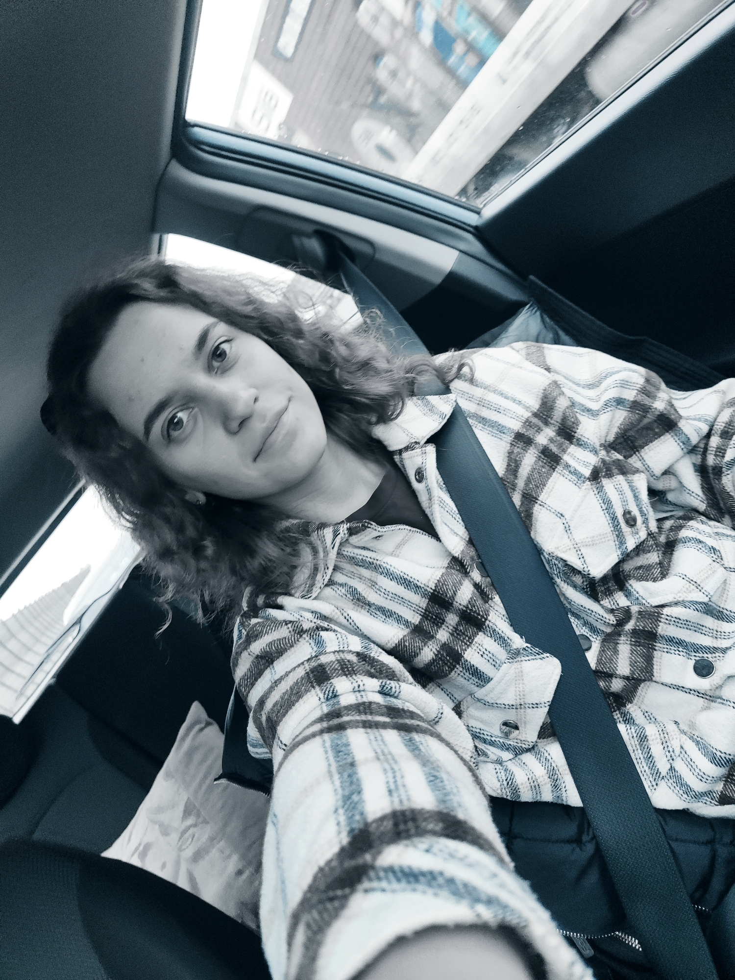 A young woman is depicted in a selfie, taken from a slightly low angle, inside a car. She has dark, curly, shoulder-length hair and looks directly at the camera with a gentle, slightly smiling expression. She is dressed in a plaid flannel shirt, featuring a pattern of white, blue, and dark grey or black checks, worn over a dark undershirt. A dark blue seatbelt is prominently fastened across her chest, securing her in the vehicle.
The interior of the car is visible, featuring dark-colored seats and headrests, and a light-colored cushion behind her. Through the car's windows, a blurred outdoor scene suggests buildings or urban structures, indicating a city or suburban setting. The bright light filtering through the windows confirms it is daytime. The image has a somewhat cool, desaturated color palette. No discernible text is visible anywhere in the frame. The overall impression is a casual moment captured during travel or while waiting in a car.