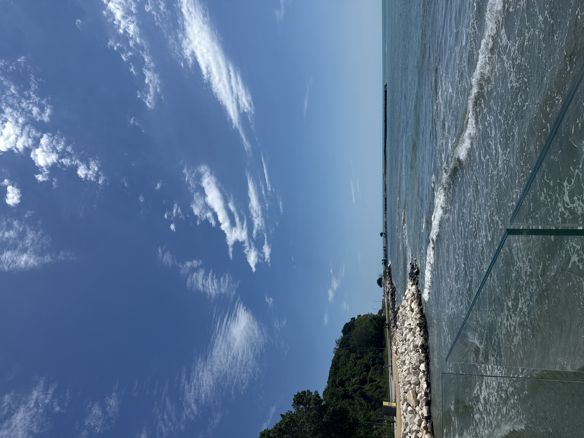 This media file captures a vibrant daytime scene along a coastal area under a bright blue sky. The image primarily features the expansive sky, the sea, and a section of the shoreline.
The sky, occupying a large portion of the frame, displays a spectrum of blue hues, from a deep, rich cobalt on the far left to a lighter, hazy azure near the horizon on the right. It is dotted with scattered white, wispy clouds (cirrus) and some smaller cumulus fragments, stretching horizontally across the upper part of the scene.
The sea, a dynamic blue-green, occupies the right side of the image, meeting the sky at a clear horizon line. Gentle, white-capped waves are visible, particularly in the foreground and near the shore, indicating an active water surface. A long, dark, horizontal structure, possibly a breakwater or pier, extends into the distant water, marking the transition to calmer waters further out.
The land element runs along the bottom of the image, featuring a lush, densely forested hillside with dark green trees and foliage, suggesting a natural park or cliffside. Below this greenery, a protective barrier of light-colored riprap (large rocks) lines the edge of the water. Adjacent to the rocky shore is a paved pathway or promenade, with faint hints of small structures like streetlights or signage in the distance. In the immediate foreground on the right, a modern, transparent barrier, likely glass or plexiglass, partially frames the view of the water, suggesting a public viewing area.
The scene conveys a peaceful and serene atmosphere, bathed in bright sunlight, suggesting late morning or early afternoon on a clear day. No people or visible text are present in the image. The setting aligns with a beautiful coastal environment, consistent with Varna, Bulgaria.