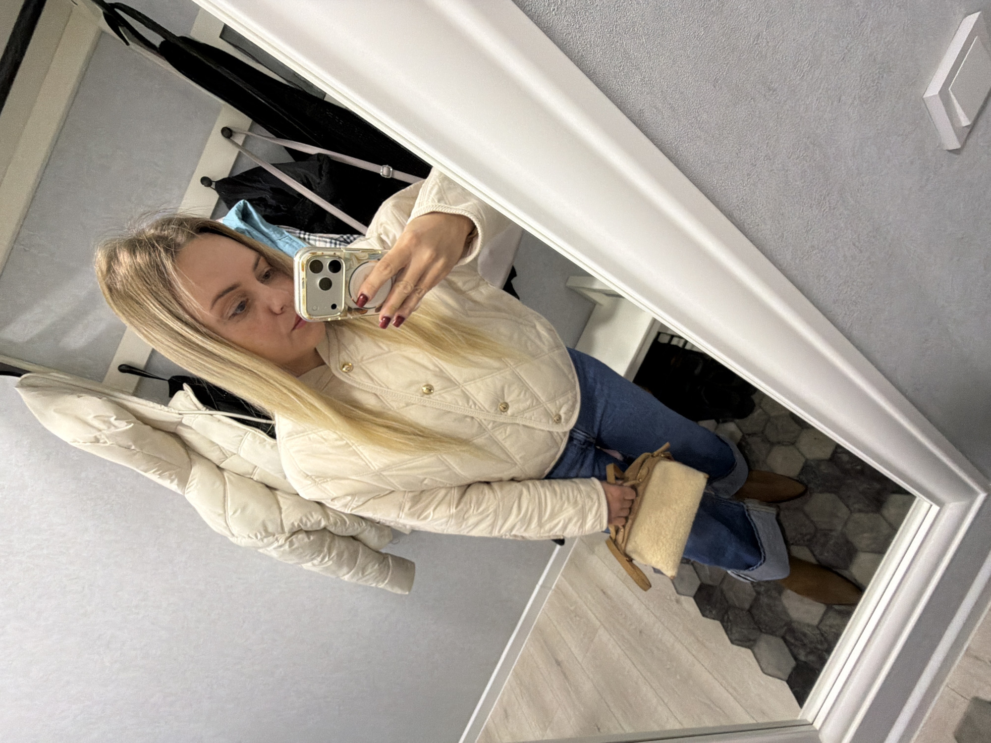 A woman stands indoors, taking a selfie in a mirror. The woman has long blonde hair and is wearing a cream-colored quilted jacket, blue jeans, and brown shoes. She is holding a smartphone with a gold case in her right hand and a cream-colored handbag in her left. The reflection in the mirror also shows various items in the background, including coats hanging on a coat rack and a second jacket hanging in what appears to be a closet space. The floor is tiled with a hexagonal pattern. The background wall is gray. There is no evidence to suggest this photo was taken in Votkinsk, Russia.
