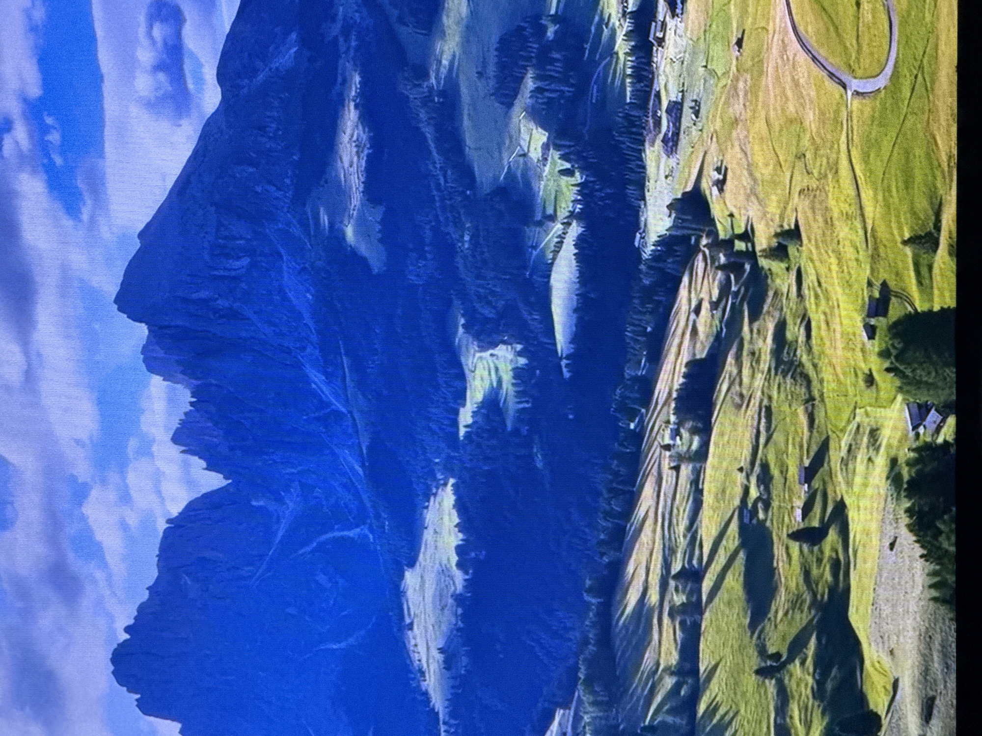 This media file captures a dramatic aerial view of a mountainous landscape bathed in the warm, golden light of a late afternoon or early morning. The scene is dominated by a colossal, dark blue mountain that dominates the left side of the frame. Its sheer, imposing cliffs create a stark contrast with the vibrant, sun-drenched green pastures stretching out on the right.
The mountainous terrain is deeply etched with ravines and shadowed valleys, suggesting a rugged and ancient geological formation. Scattered across the green hills are small, dark structures, possibly homes or farm buildings, nestled amongst the trees. The slopes are intricately patterned with the long shadows cast by the setting or rising sun, highlighting the undulating contours of the land.
A prominent feature in the upper right of the image is a paved, circular road or path, hinting at a public space or recreational area within this natural setting. The sky above the mountain is a brilliant blue, dotted with soft, white clouds that appear somewhat textured, suggesting a slightly hazy or diffused light.
There is no visible text in the image. The overall impression is one of awe-inspiring natural beauty, with the dramatic interplay of light and shadow emphasizing the scale and grandeur of the mountainous region. The location context of Dubai, United Arab Emirates, is not visually apparent in the provided image.