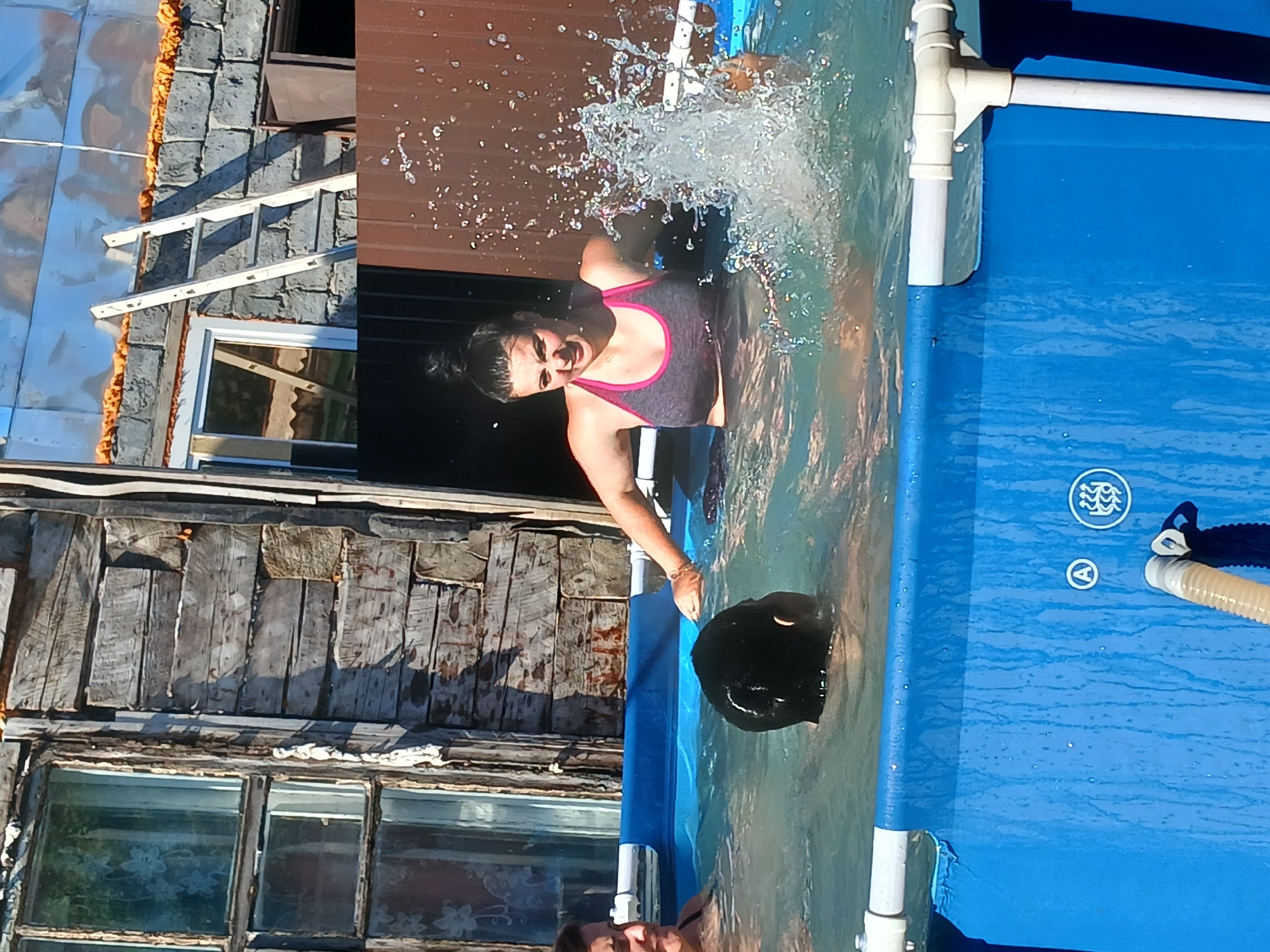 This media file shows an outdoor scene with a blue inflatable pool filled with water. Two individuals are partially visible in the pool. One person, a woman with dark hair pulled back, is in the foreground and appears to be splashing water. She is wearing a dark tank top with pink trim. Her expression suggests exertion or enjoyment. Her arm is extended, and water droplets are frozen mid-air around her, indicating movement.
Behind her, another person's head, appearing to be dark and wet, is submerged in the water, with only the top of their head visible. The pool has white PVC piping supporting its structure, and some of this piping is visible at the edge of the frame. The water is clear and reflects the surroundings, with small ripples on the surface.
The background is a cluttered outdoor environment. To the left, there is a weathered wooden structure with clapboard siding and visible nails. Above this, a section of a building with a corrugated metal roof and a white ladder leaning against a stone-like facade is visible. There is also a window with a wooden frame and glass panes reflecting light. The overall impression of the setting is rustic and possibly rural. The time of day appears to be sunny, with strong shadows cast by the structures. The weather seems warm enough for swimming. There is no visible text in the media file.