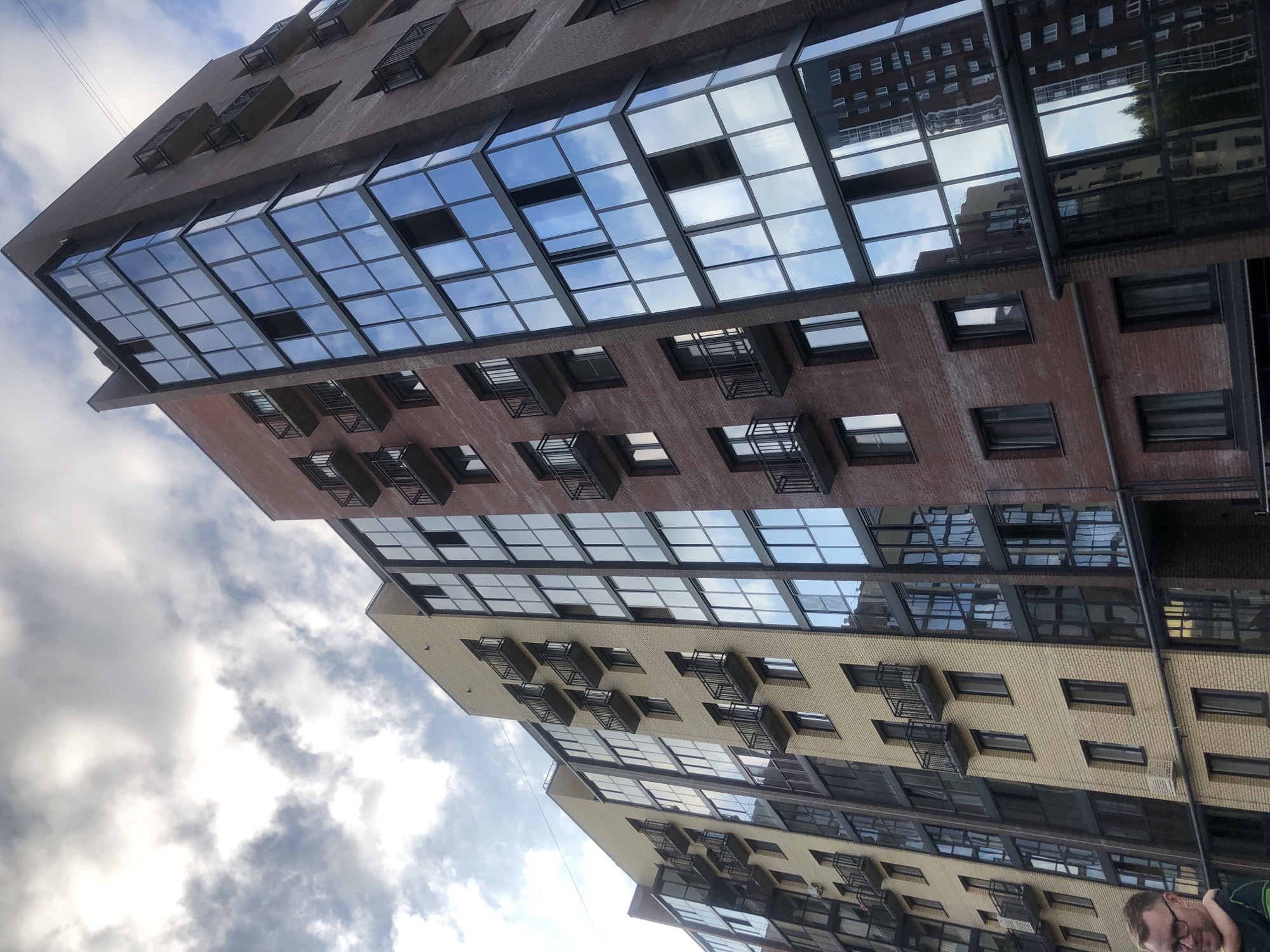 The image shows a modern apartment building with a brick facade and large glass windows reflecting the cloudy sky. The building has multiple balconies with black railings. The perspective is from a low angle, looking up at the building. A portion of a person's face and shoulder is visible in the lower left corner of the image. The surrounding environment appears to be urban, with other buildings visible in the reflection of the glass. The overall scene suggests a residential area in Zhukovo, Russia, characterized by contemporary architecture.