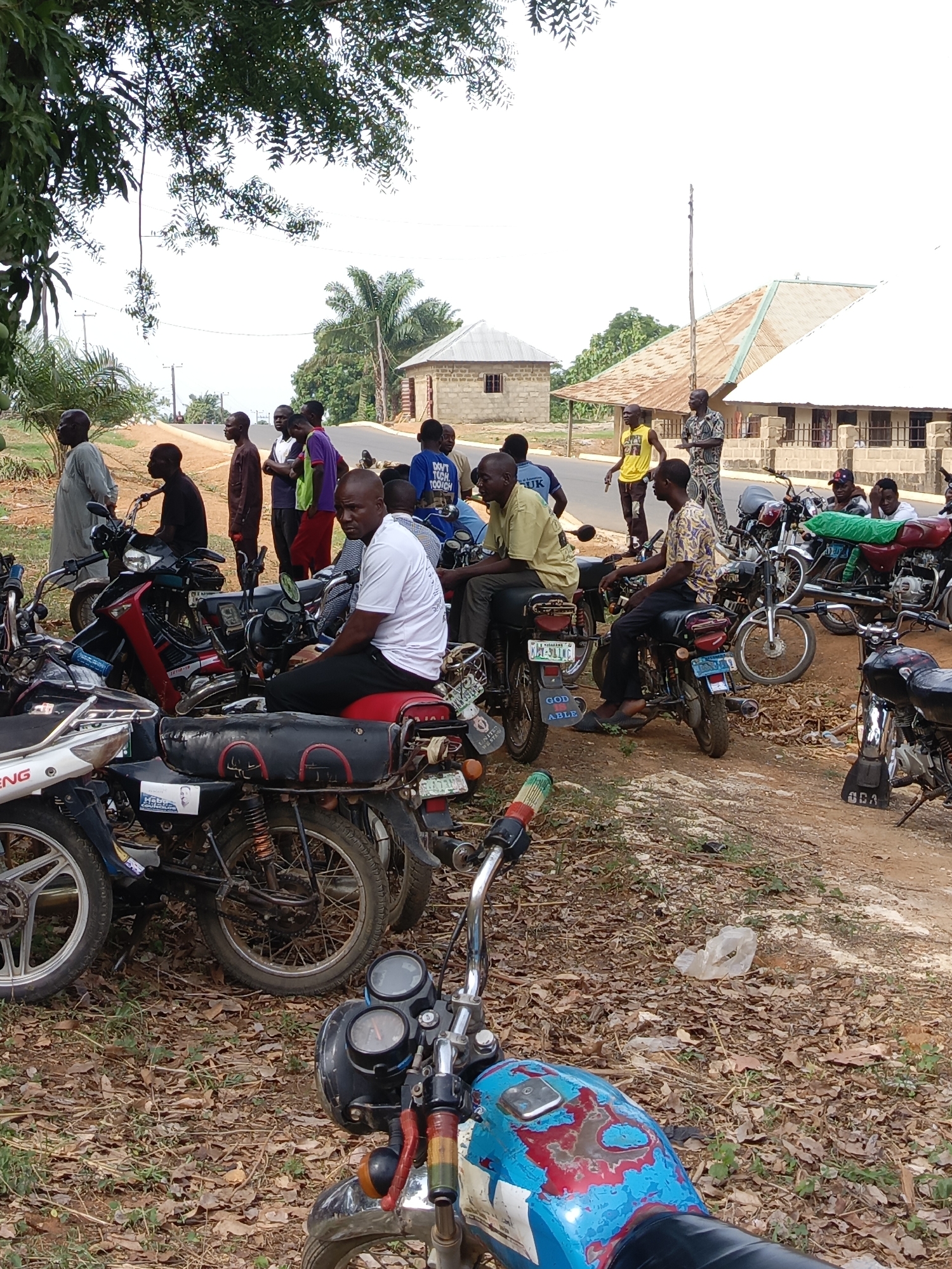 This media file captures an outdoor scene in what appears to be a rural or semi-urban area in Andaha, Nigeria, characterized by dry, leafy ground and simple architecture. The foreground and midground are dominated by numerous men and motorcycles, suggesting a gathering point, possibly a motorcycle taxi stand or a community space.
A large group of men, mostly young to middle-aged, are scattered throughout the image. Some are seated on their motorcycles, appearing to wait or rest, while others stand in small groups, seemingly conversing or observing. One man in a white t-shirt and black trousers, sitting on a red motorcycle in the midground, looks directly at the camera with a neutral expression. Another man in a light green shirt and light-colored trousers sits on a motorcycle further left, looking away. In the background, several men stand by the roadside.
The motorcycles are diverse in model and condition, typical of those used for transport in the region. Many are parked closely together. In the background, beyond the road, a few single-story buildings with corrugated metal roofs are visible, set amidst trees and foliage. The sky is bright, indicating daytime, likely morning or afternoon, with no harsh shadows. The overall atmosphere seems casual and unhurried.
Visible text includes: "DON'T TOUCH TOO MUCH" on a blue t-shirt, "GOD ABLE" on a motorcycle's mudguard, and license plates like "KM-511HG". A small, indistinct image of a face (possibly political) is seen on a sticker on one motorcycle. Other partial texts like "OK" on a yellow shirt, "SUK" on a blue shirt, and "SDA" on a motorcycle part are also present.