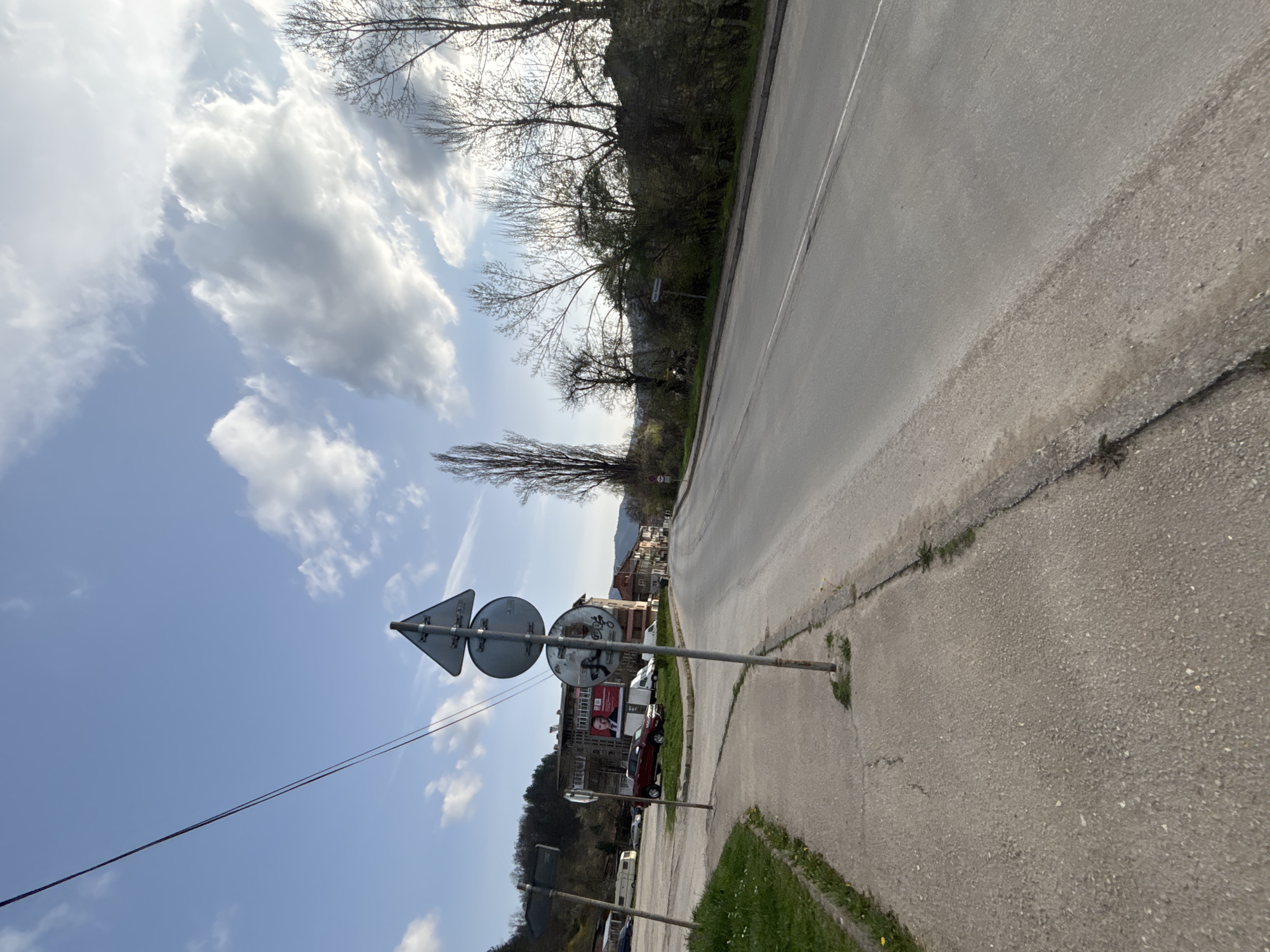 The image depicts a street scene in Troyan, Bulgaria. The view is from a sidewalk with a combination of asphalt and concrete textures. A metal signpost displaying traffic signs (a yield sign and a roundabout sign) is visible in the lower left foreground, oriented at an angle towards the sky. Utility wires run parallel to the street, extending towards the background.
The street stretches into the distance, bordered by a tree-lined area on the right. Buildings, including some with red roofs, and parked cars are visible on the left side of the street. A billboard is also visible among the buildings.
The sky is blue with scattered white clouds. The lighting indicates it is daytime.