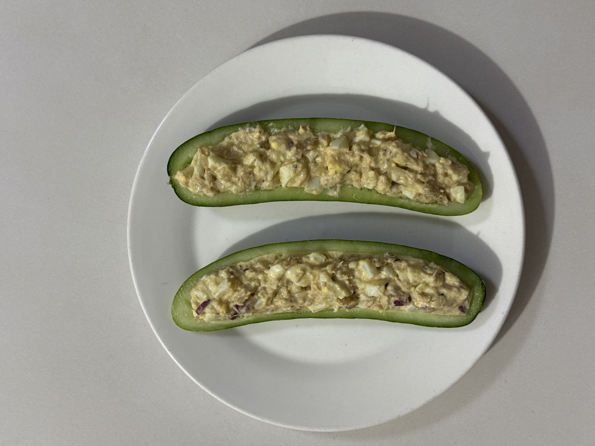 Two halves of a cucumber, hollowed out and filled with a tuna salad mixture, rest on a white circular plate. The tuna salad appears to contain chopped hard-boiled eggs and red onion. The plate is positioned on a neutral-colored surface, possibly a countertop, with a subtle shadow cast to the right of the plate. The scene is lit from above, creating soft lighting. This image is a close-up, top-down view of the food, with no discernible urban or environmental context suggestive of Davao City, Philippines, or any specific location.