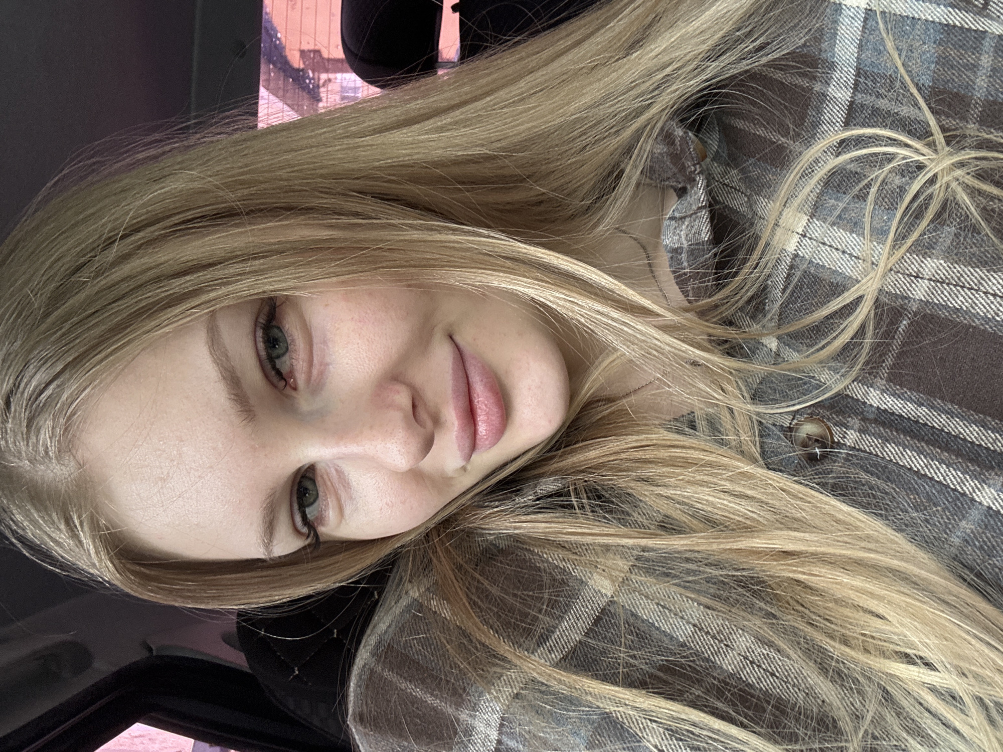 A young woman with blonde hair is photographed indoors, possibly in a car. Her face is angled slightly, revealing her features. She is wearing a brown and grey plaid shirt. In the background, a blurry glimpse of a building and possibly other objects is visible through a window. There is no concrete information to place the subject in the city of Perm, Russia.