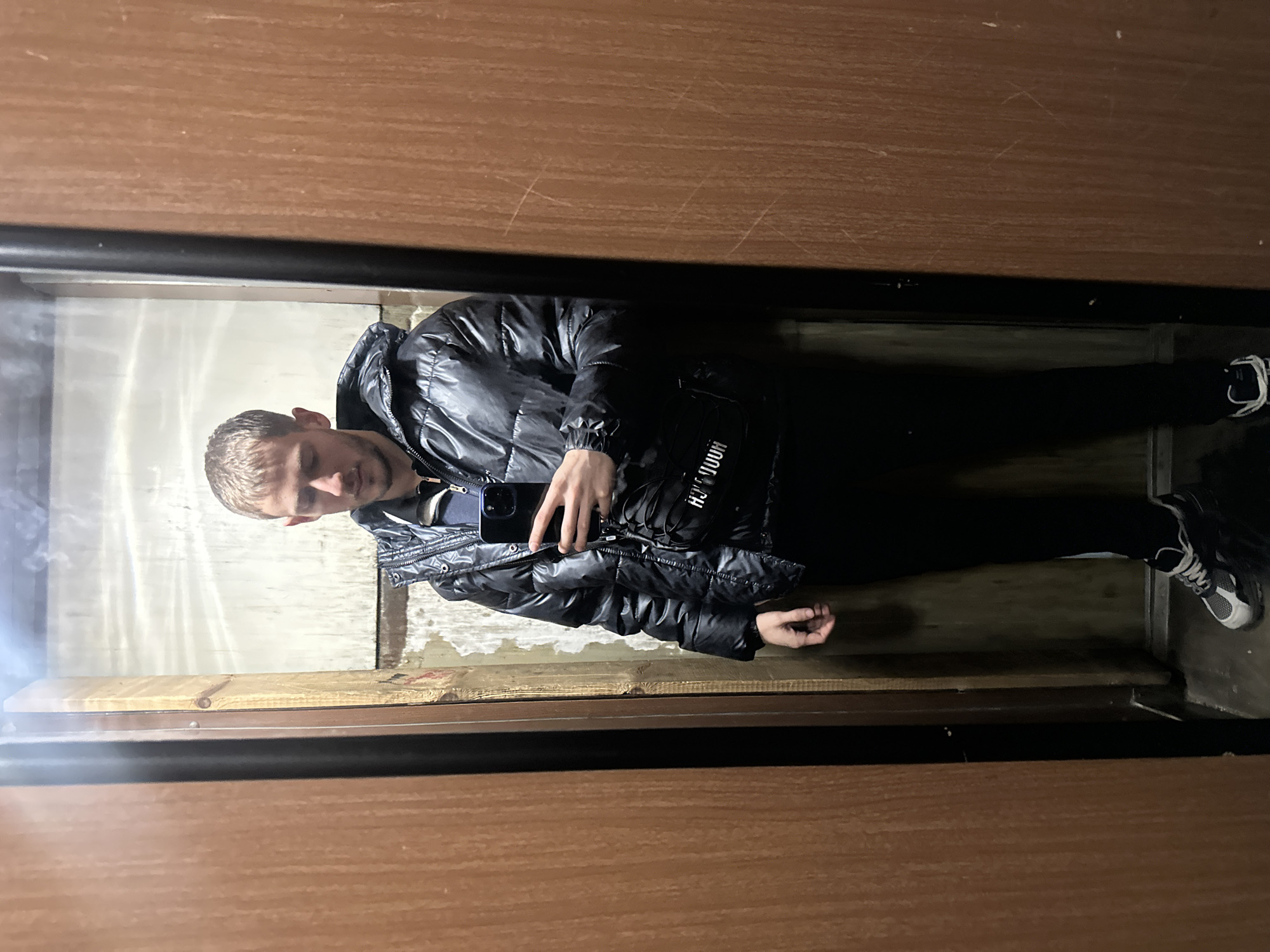 A man is taking a photo of himself in a mirror. He is wearing a black puffer jacket, black pants, and black and white sneakers. He is holding a smartphone in his left hand. The mirror is framed with black trim and mounted on a wall with brown wood paneling. The floor appears to be stained concrete. The location is inside an elevator, and it is not possible to determine the city without additional information.
