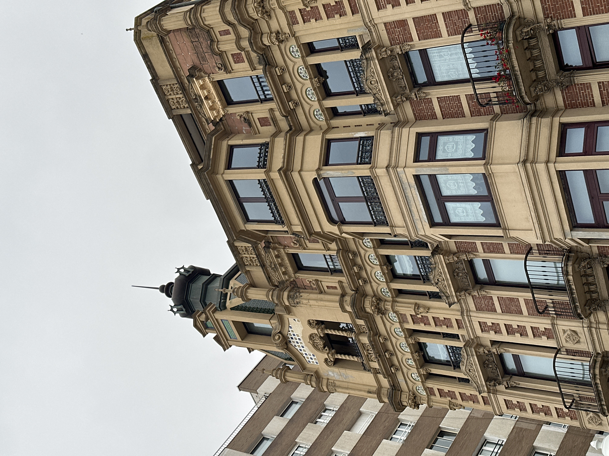 The image shows a building in Gijón/Xixón, Spain. The building has a distinctive architectural style, featuring ornate detailing, including carvings, balconies with metal railings, and a tower with a pointed spire. The facade is a mix of red brick and beige-colored decorative elements. Windows are present on multiple floors. The sky is overcast. A more modern building with striped, alternating light and dark horizontal patterns can be seen below the ornate building.