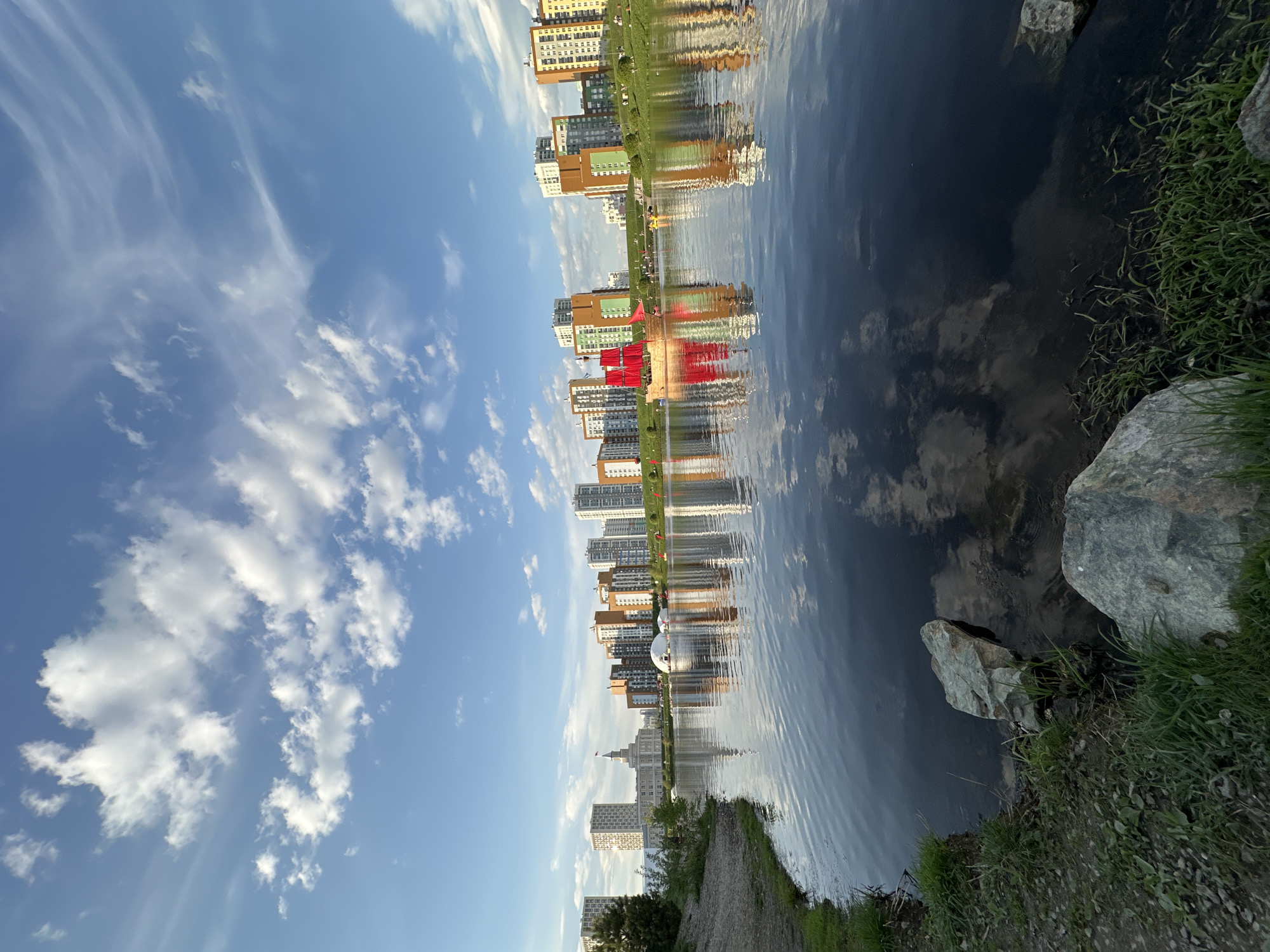 The image depicts a serene urban landscape in Yekaterinburg, Russia. A body of water reflects the sky with scattered clouds and the city's architecture. Buildings of varying heights and colors, including a prominent red structure and a domed building, line the water's edge. The shoreline is defined by rocks and patches of grass. A narrow path runs along the shore. The composition is inverted, with the sky appearing at the bottom and the water at the top.