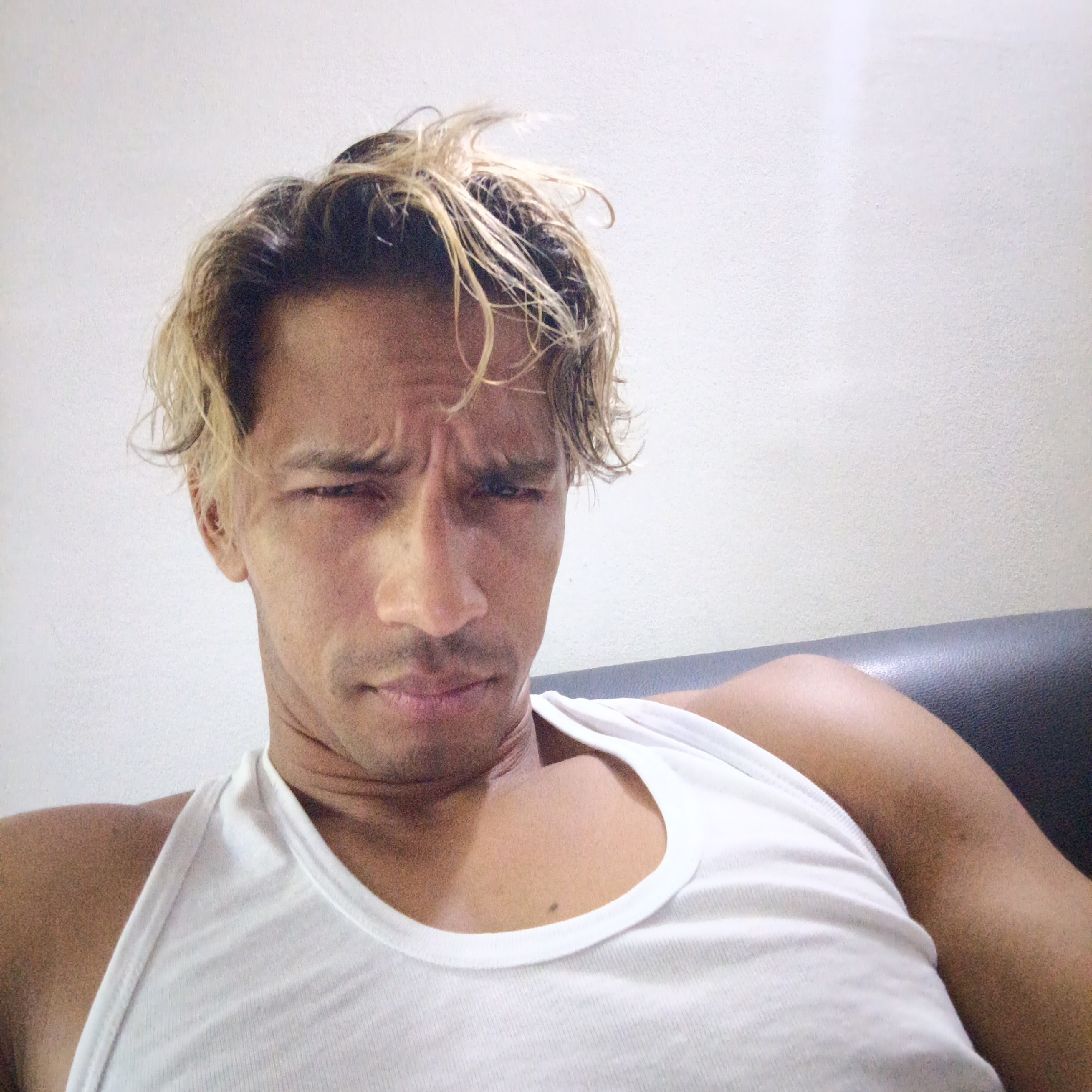 A close-up, forward-facing selfie captures a man with tanned skin and tousled blonde-streaked hair. He is wearing a white tank top and has a furrowed brow, suggesting a questioning or displeased expression. The background is a plain white wall, and a dark surface, possibly a couch, is visible in the lower right corner. The image was taken indoors. The location is Piloncito, Venezuela.