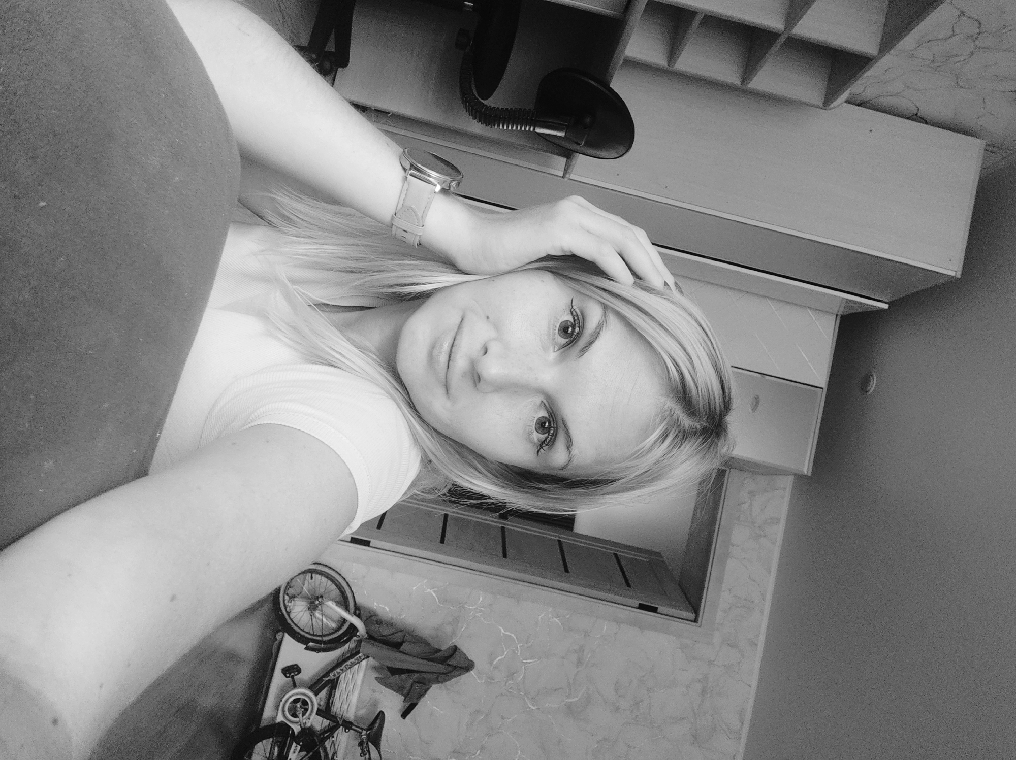 The image is a black and white, medium shot selfie taken indoors. A young Caucasian woman with blonde hair, wearing a white t-shirt and a watch on her left wrist, is the main subject. Her right hand is resting on her head. She is positioned in the foreground, looking towards the camera with a neutral expression.
The background reveals a room with furniture including a shelving unit, a desk chair, and a closet. A doorway is visible, leading to another area. To the right of the doorway, a bicycle is leaning against the wall. The walls appear to have a textured wallpaper pattern. The floor is made of wood. The lighting suggests an indoor setting, possibly a residential room. The specific location in the city of Sterlitamak, Russia, cannot be determined from the visual information provided in the image.