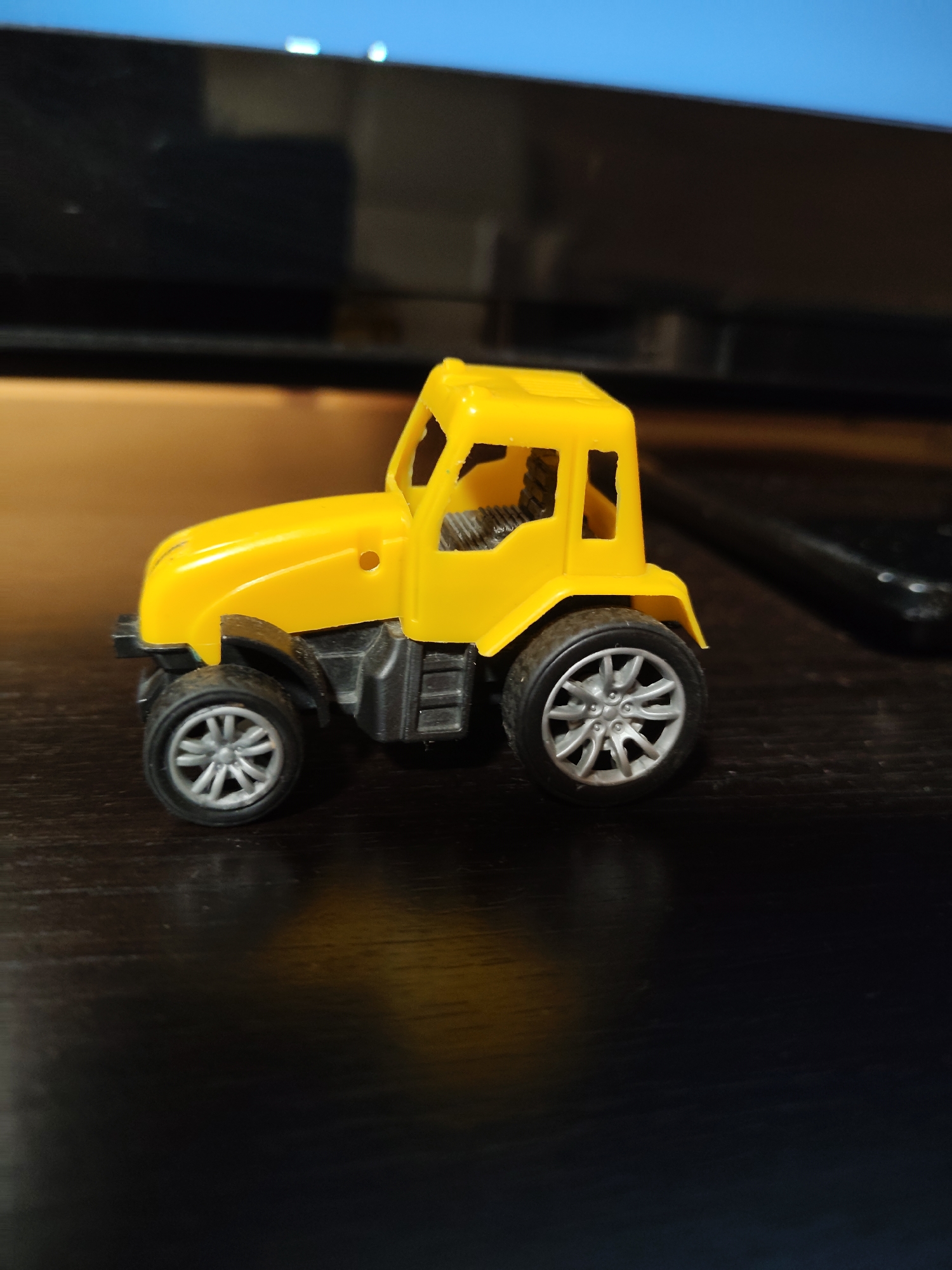 A yellow toy tractor is positioned on a dark, reflective surface, likely a table. The tractor has a detailed design, featuring a yellow cab with visible interior textures, black molded plastic wheel wells, and silver-colored wheels with a multi-spoke design. The surface it rests on shows a faint reflection of the tractor and a warm, yellowish light source, suggesting indoor lighting. In the background, a dark, possibly electronic device, is partially visible, and above that, a blurry blue area. The image appears to be a close-up shot, focusing on the toy. The location of Фестивальное Снт, Russia, is not evident in the provided image.