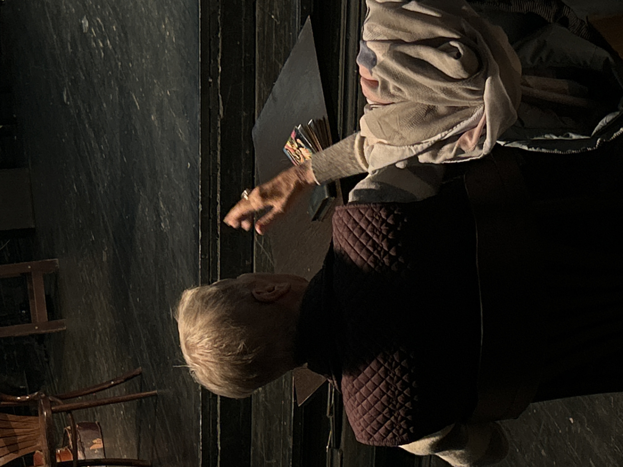 A person with short, light hair sits in a chair covered with a beige blanket. The person wears a burgundy quilted jacket and their hand is extended toward a small stack of cards. The setting appears to be indoors, with a wooden floor and dark walls, and a wooden chair nearby. The location is unknown.