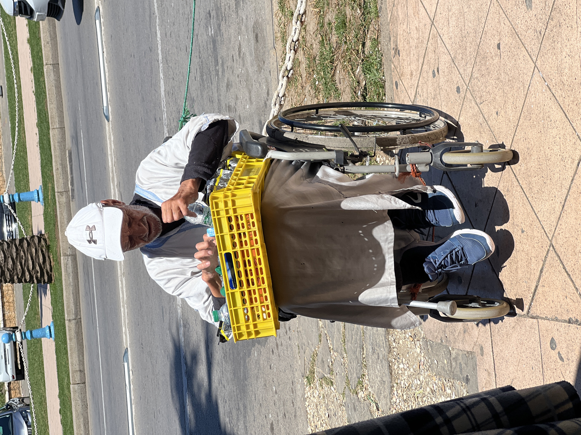 The image shows an elderly man in a wheelchair holding a crate of water bottles. He wears a white baseball cap and is positioned on a sidewalk next to a street. The location is M'Diq, Morocco.