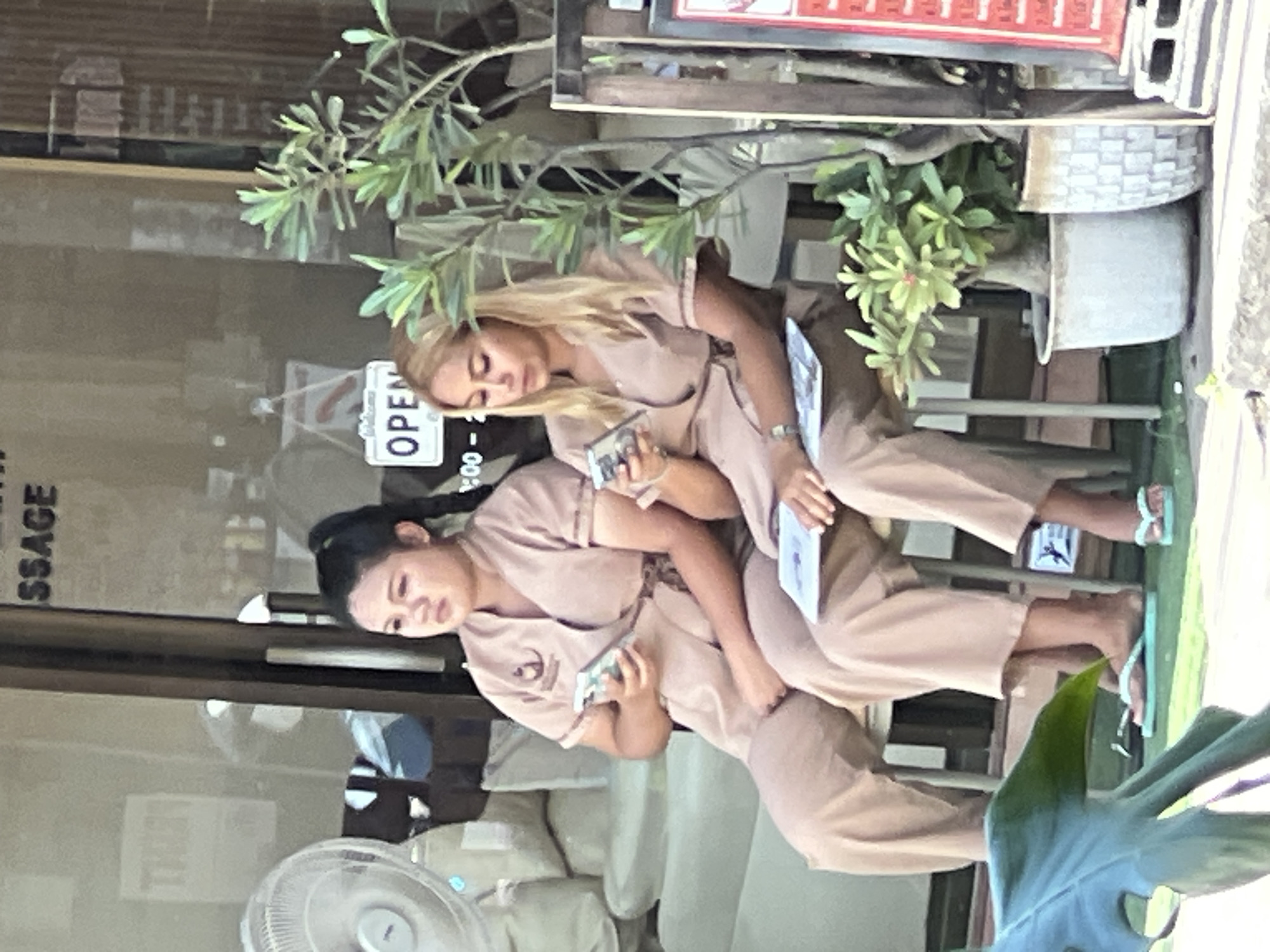 Two women, appearing to be of Thai nationality, are seated on chairs outside a storefront in Ko Samui District, Thailand. The storefront has a large glass window with text on it, partially obscured, but "SSAGE" is visible. A white sign with "OPEN" is also visible through the window. Both women are wearing similar light brown, wrap-style uniforms. The woman on the left has her hair in a ponytail and is holding a smartphone. The woman on the right has blonde hair and is also holding a smartphone and a booklet. There is a potted plant to the right of the women. In the foreground, a large green leaf is visible. The overall environment suggests a business setting, possibly a spa or massage parlor, given the visible text.