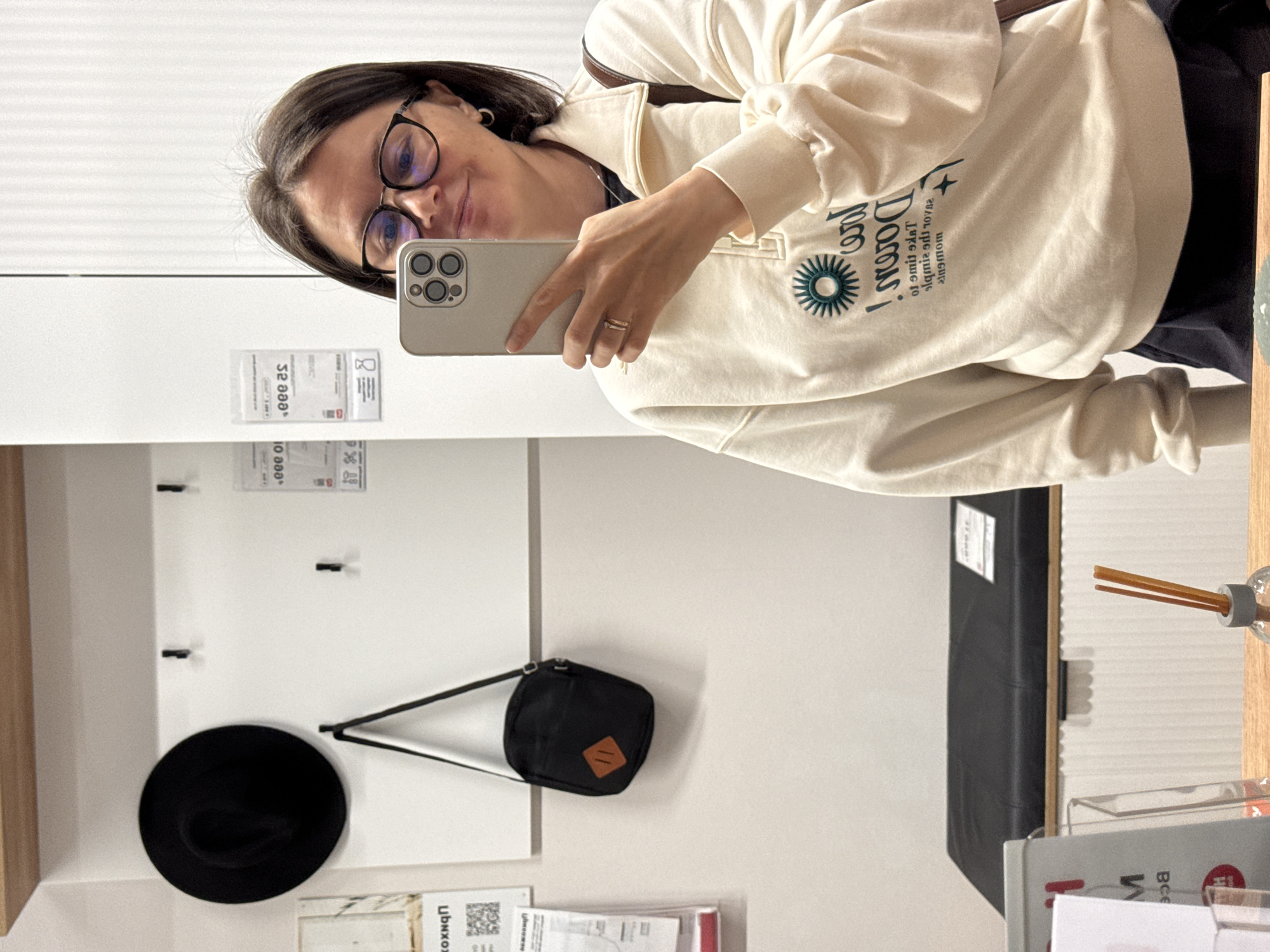A woman is taking a selfie with a smartphone in what appears to be a showroom or possibly a home setting. The background suggests a retail or display environment with various objects and furniture.
The woman is wearing glasses and a light-colored sweater with writing and an emblem on the front. She is holding the smartphone at arm's length.
In the background, there is a white wall with hooks, upon which hang a black hat and a small black bag. Additionally, there are papers and tags affixed to the wall. A dark upholstered furniture piece is visible to the right, along with what appears to be a wooden surface and a glass container with sticks, suggesting an aroma diffuser.
The location in the city of Izhevsk, Russia is not apparent from the image.