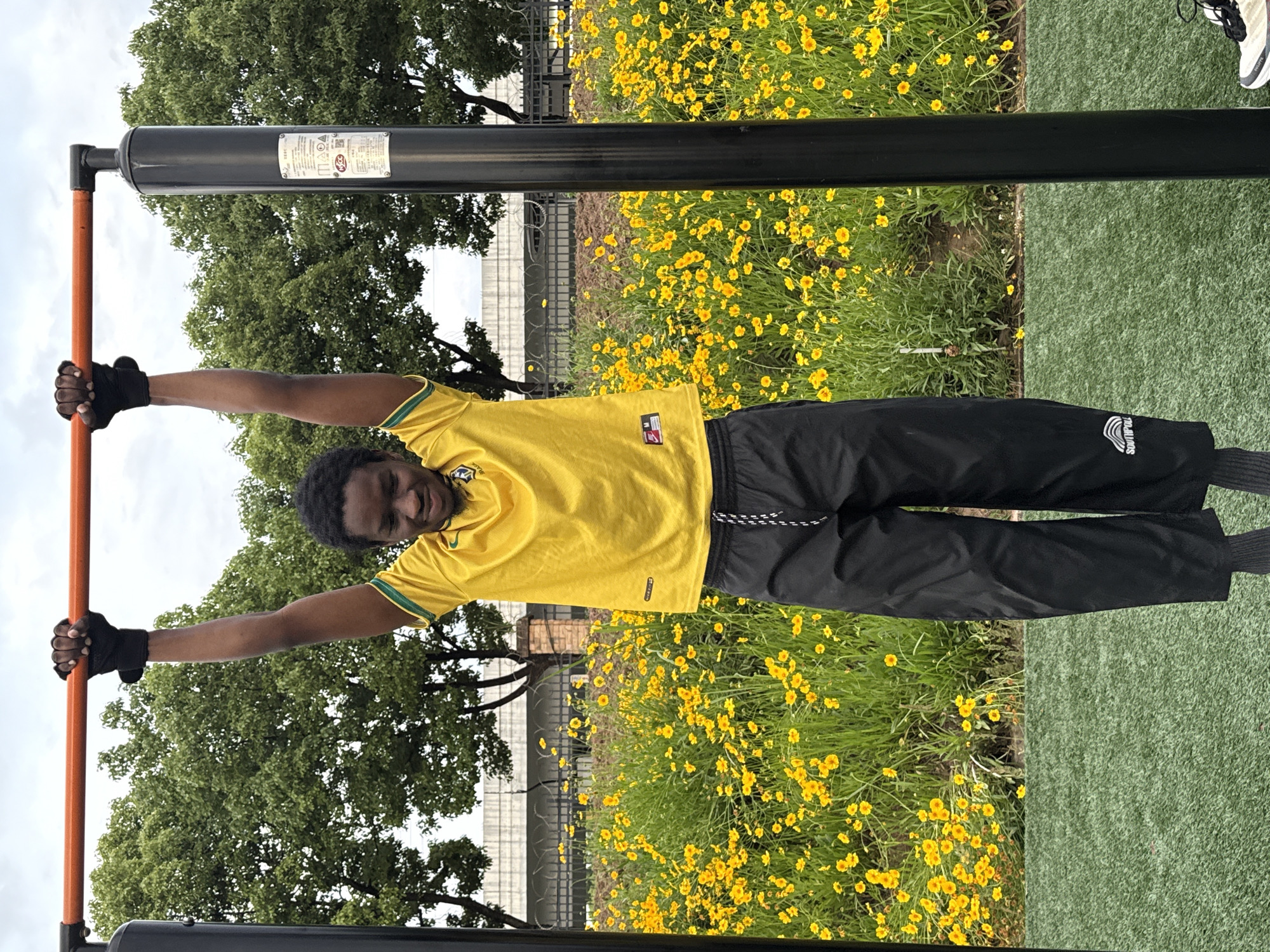A man hangs horizontally from a pull-up bar in an outdoor setting. He wears a yellow soccer jersey and black athletic pants with black wrist wraps. The pull-up bar is situated in a park-like environment with green grass and yellow flowers. The background includes trees and a partially visible building. The location is Jiangjin, China.