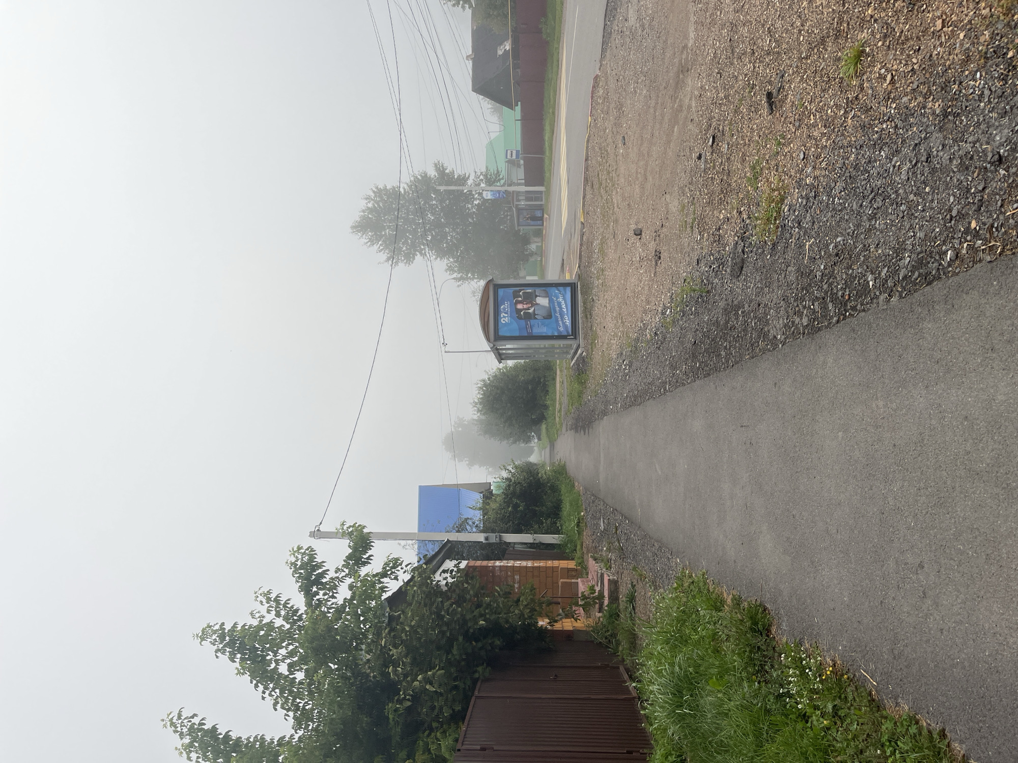 The image depicts a street scene in Blagoveshchensk, Russia, characterized by dense fog. A paved sidewalk runs along the left side of the image, bordered by grass and a brown metal fence. Adjacent to the sidewalk is a house with orange brick lower walls and a white upper story, with a small porch. Further down the street, a house with a blue roof is visible. On the right side of the street, there is a bus stop shelter with an advertisement. Across the road, houses and trees are partially obscured by the fog. Electrical wires are strung overhead. The ground along the road is covered in gravel and dirt. The overall atmosphere is overcast and quiet, suggesting a low-visibility day.