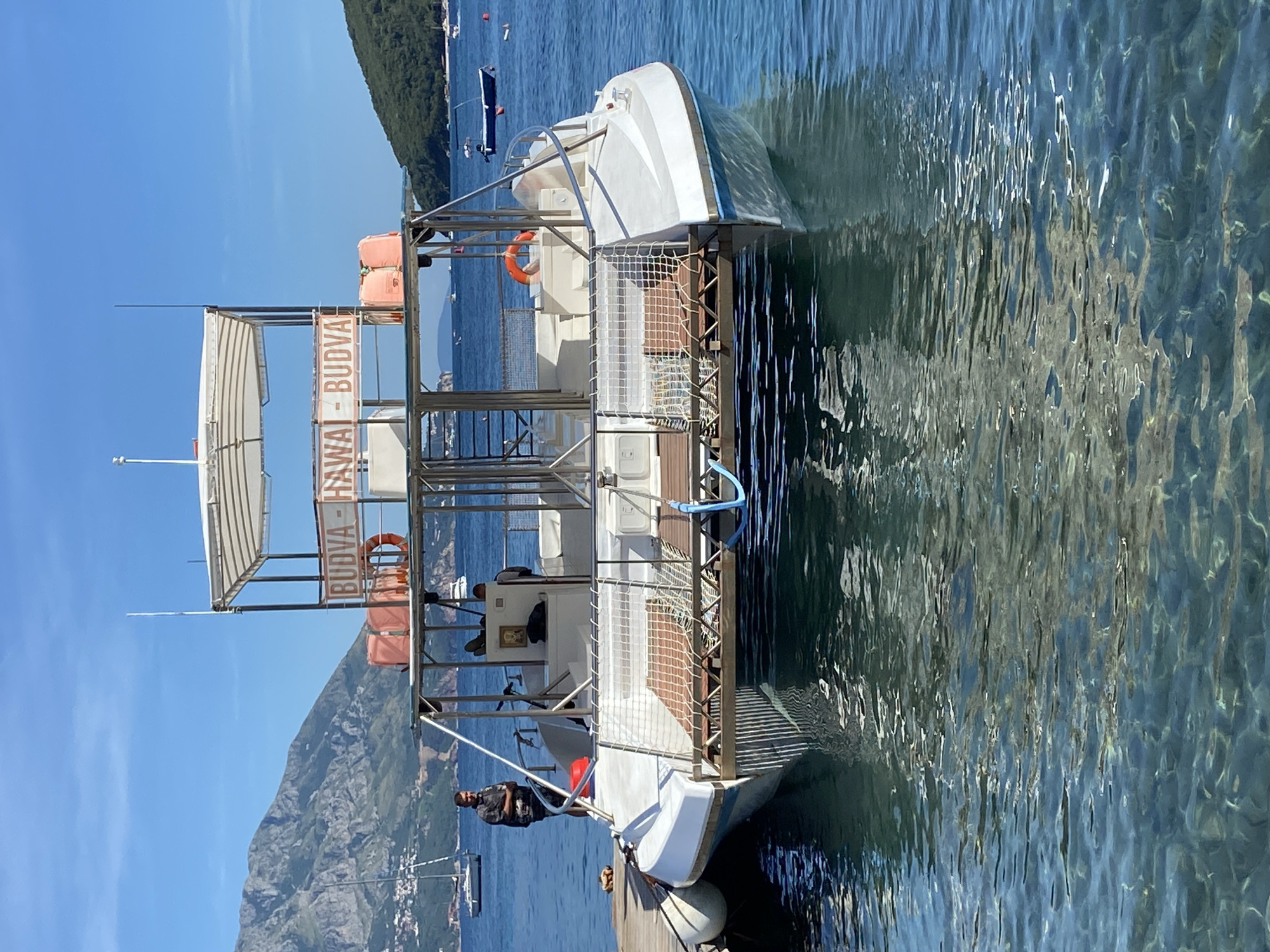 A white catamaran is docked at a pier in the clear blue waters of Budva, Montenegro. Mountains, some covered in green vegetation and others rocky, form the backdrop. Several sailboats and smaller boats are visible in the distance. The catamaran has a bimini top providing shade over a deck area with seating. An orange life preserver and a life ring are visible on the deck. A sign on the upper structure reads "BUDVA - HAWAI - BUDVA". Two men are on the boat; one is standing near the stern on the pier, and another appears to be in the cabin area. The water is calm with a visible reflection of the boat and the surrounding environment. The pier is made of wood.