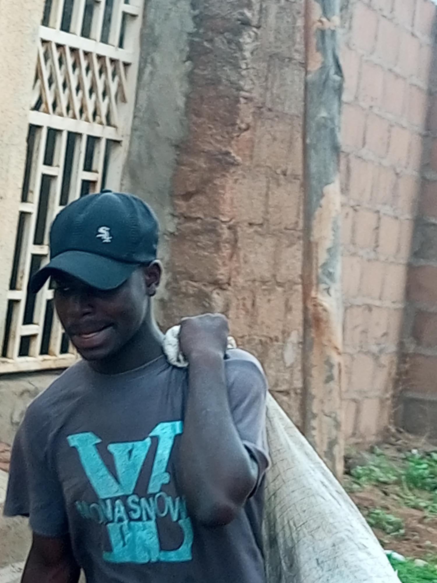 A young man wearing a black baseball cap with a white logo and a dark gray t-shirt with a large light blue "V" and the text "NOVASNOV" below it, is carrying a large white sack over his right shoulder. He is looking downwards and smiling slightly. He is standing in front of a rough, light-colored wall with a vertical textured band in the center, and to his left, there is a white barred window. The ground is covered with some green vegetation.