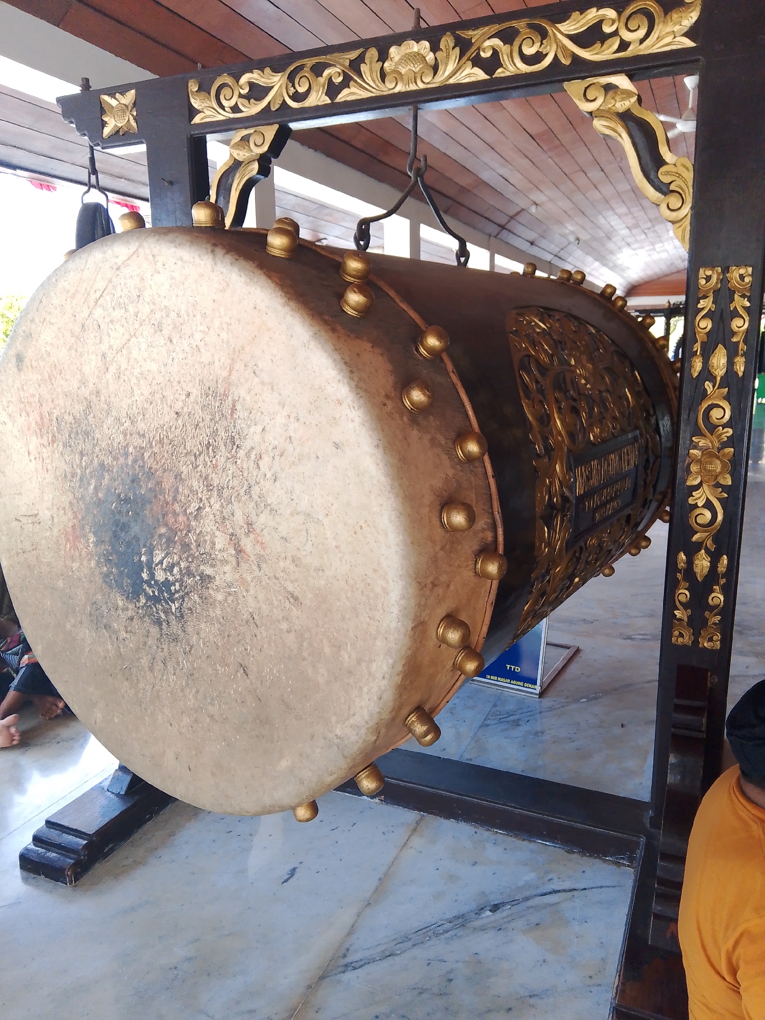 A large, traditional drum, likely a bedug, is displayed on a dark wooden stand with ornate gold carvings. The drum itself has a light-colored head with a dark, mottled area in the center, possibly from use or repair. The body of the drum is dark brown with decorative gold carvings and numerous small gold studs around its circumference. The stand is elaborately decorated with gold scrollwork and floral motifs, and it is positioned on a marble floor. In the background, a wooden ceiling is visible, with some sunlight filtering through. Partially visible at the bottom of the frame are the legs of a person and a section of an orange shirt, suggesting the presence of onlookers. A blue sign with the text "TTD TA'MIR MASJID AGUNG DEMAK" is visible near the drum, indicating its location at the Great Mosque of Demak in the city of Demak, Indonesia.