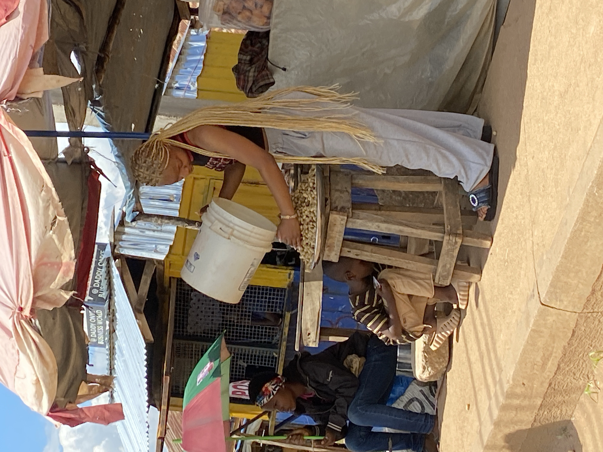 A young woman with blonde braided hair is pouring peanuts from a white bucket onto a metal tray on a wooden stand at an outdoor market stall in Bukuru, Nigeria. She is wearing a black crop top and grey sweatpants. Beside her, a young boy wearing a striped shirt and tan pants is sitting on a wooden stool, looking down. To the left, a woman in a black jacket and blue jeans sits under a pink and green umbrella, holding a small object. The market stall has a corrugated metal roof and yellow and blue walls. Bags of food are visible on a table behind the woman. The ground is sandy and there is a concrete curb in the foreground. Signs in the background read "@ DADDY ONYE BUSINESS WORLD."
