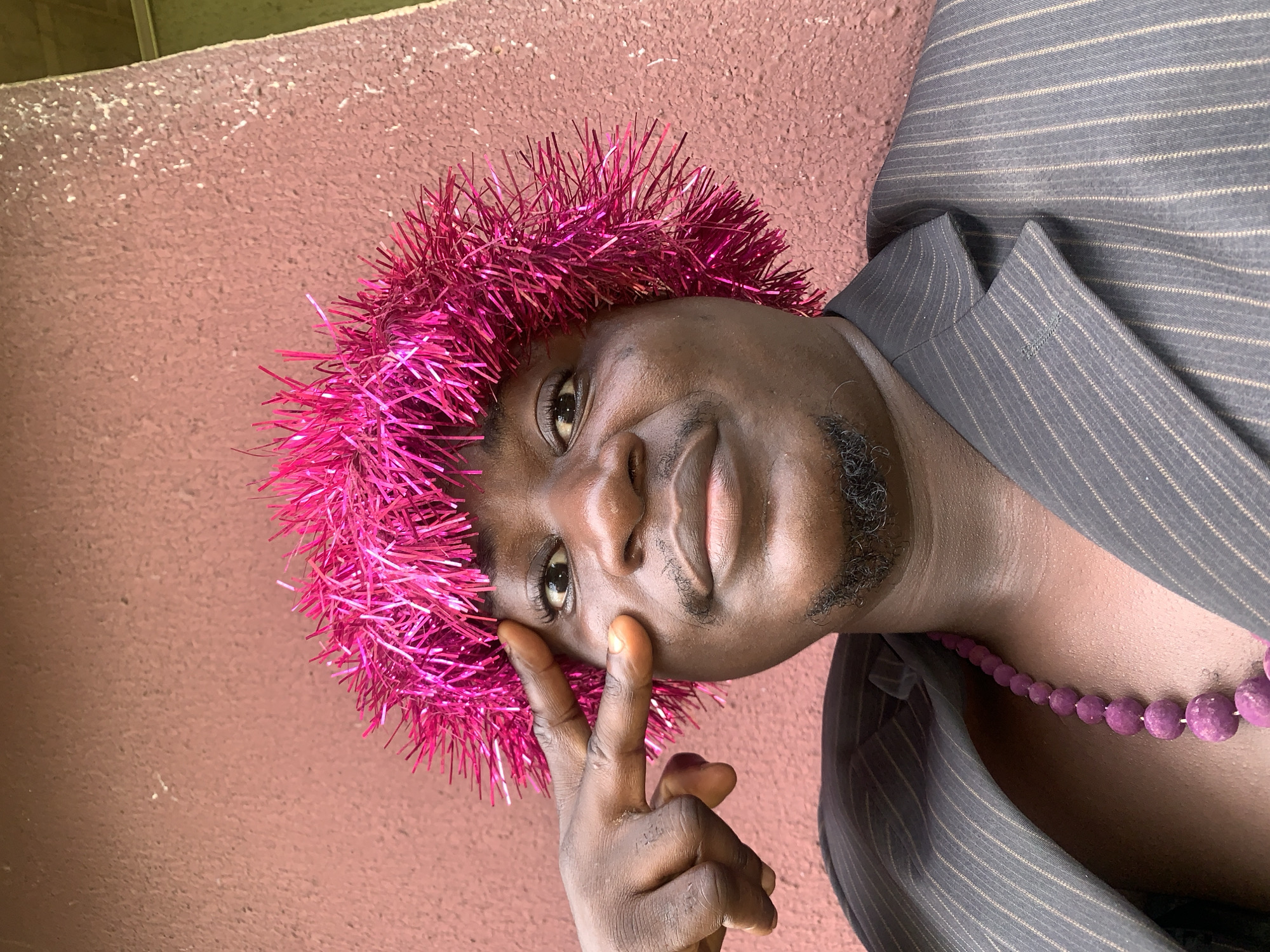 In Akwanga, Nigeria, a person with dark skin and a goatee wears a bright pink tinsel-like wig and a pinstripe suit jacket over a purple beaded necklace. They are posing for a selfie, with their left hand held up to their forehead, index finger extended. The background is a textured, reddish-brown wall.