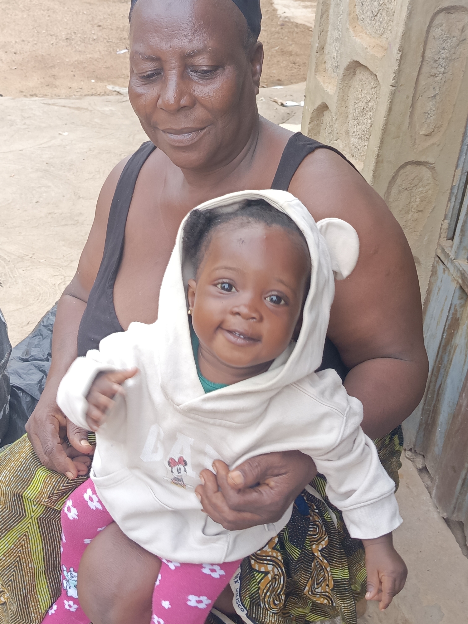 In Akwanga, Nigeria, an older woman with dark skin and a black tank top holds a baby. The baby, also with dark skin, wears a white hooded sweatshirt with Mickey Mouse graphics and pink floral leggings. The woman sits near a rough concrete wall and a rusty metal door. A patterned skirt covers her legs. A black plastic bag sits to the left. The baby smiles at the camera.