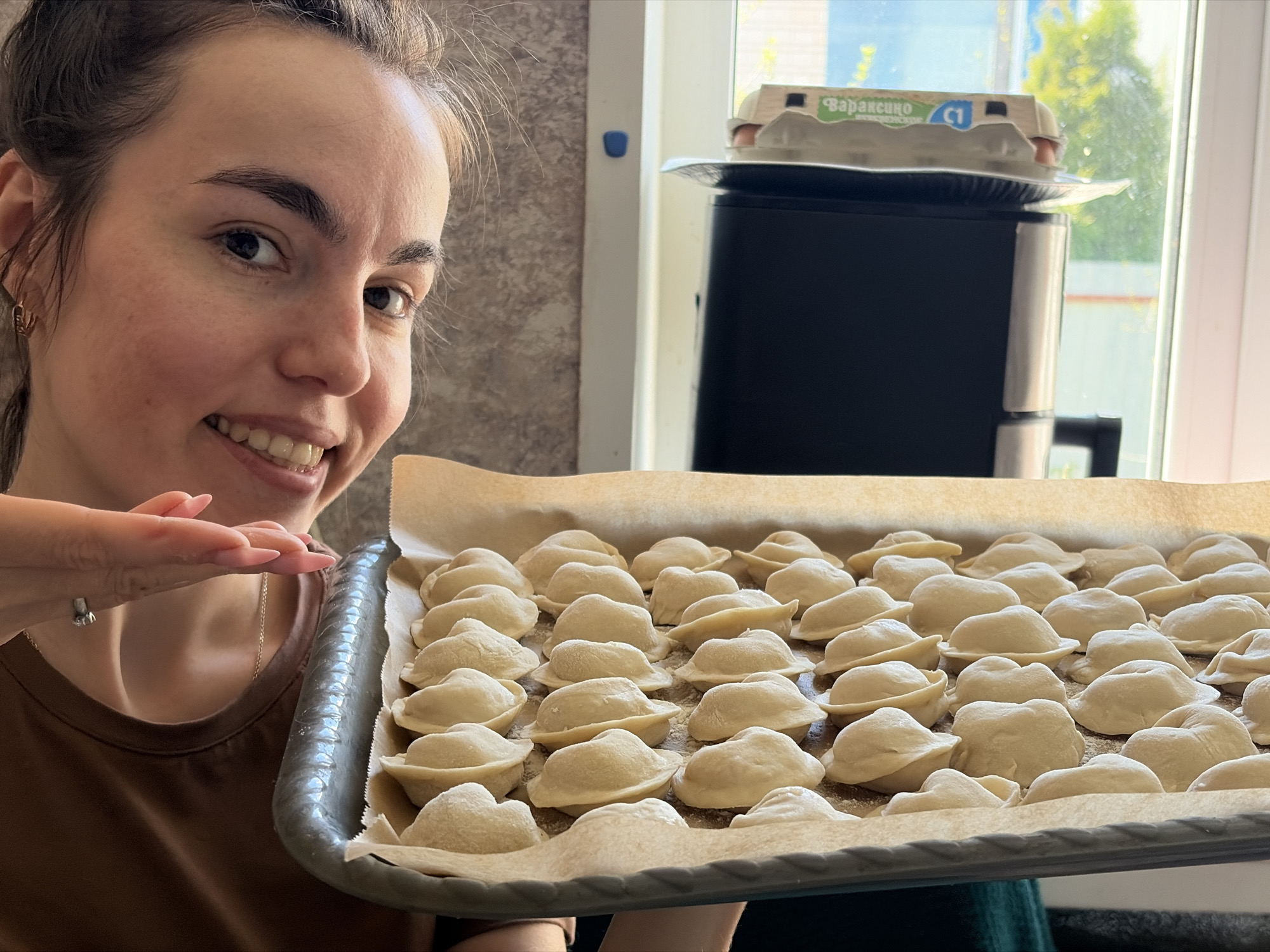 A woman is holding a baking sheet covered with rows of uncooked, dumpling-like foods. The dumplings are arranged on a sheet of parchment paper within the gray baking sheet. The woman is smiling and has her hand raised near the dumplings. In the background, an air fryer sits on a counter near a window. An egg carton with Cyrillic text is on top of the air fryer. The text on the carton suggests the location of Nizhnii Vishur, Russia.