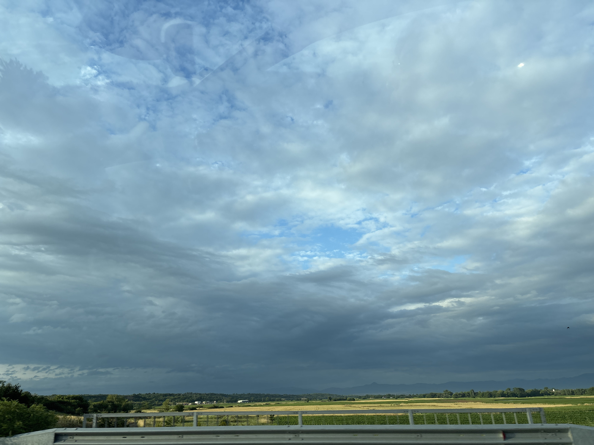 The image depicts a wide, expansive sky filled with a mixture of white and dark grey clouds, suggesting an impending storm or overcast weather. A patch of clear blue sky is visible in the upper center. The landscape below consists of agricultural fields, some appearing yellow and others green, interspersed with patches of trees. In the distance, a range of mountains can be seen, their peaks somewhat obscured by the atmospheric conditions. A metal guardrail runs horizontally across the foreground, indicating that the photograph was likely taken from a bridge or overpass. There are no people or animals visible in the scene. The image captures a rural or semi-rural environment in the vicinity of Cherna Gora, Bulgaria, characterized by agricultural land and natural topography.