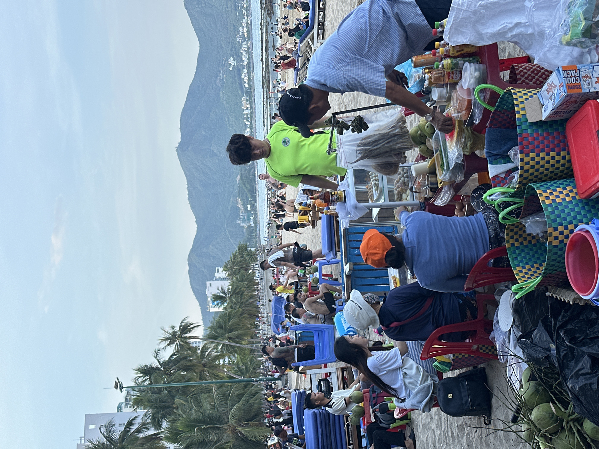 This is a photograph taken on a beach in the city of Nha Trang, Vietnam. The scene depicts a lively beachside atmosphere with numerous people enjoying the outdoors. In the foreground, a vendor stand is set up, displaying a variety of food items, beverages, and packaged goods. Two male vendors are attending to the stand, one wearing a bright green t-shirt and the other a blue checkered shirt and a black baseball cap. Several customers, including women and men, are seated at tables and chairs near the stand, some of whom appear to be consuming food or drinks.
Behind the vendor stand, the beach is populated with many beachgoers, some relaxing on lounge chairs, others walking or gathered in groups. Palm trees line the back of the beach, and in the distance, a cityscape with buildings and mountains can be seen against a hazy sky. The presence of food stalls, vendors, and a large number of people indicates a popular tourist or recreational area. The overall mood of the image is one of activity and social gathering on a beach.